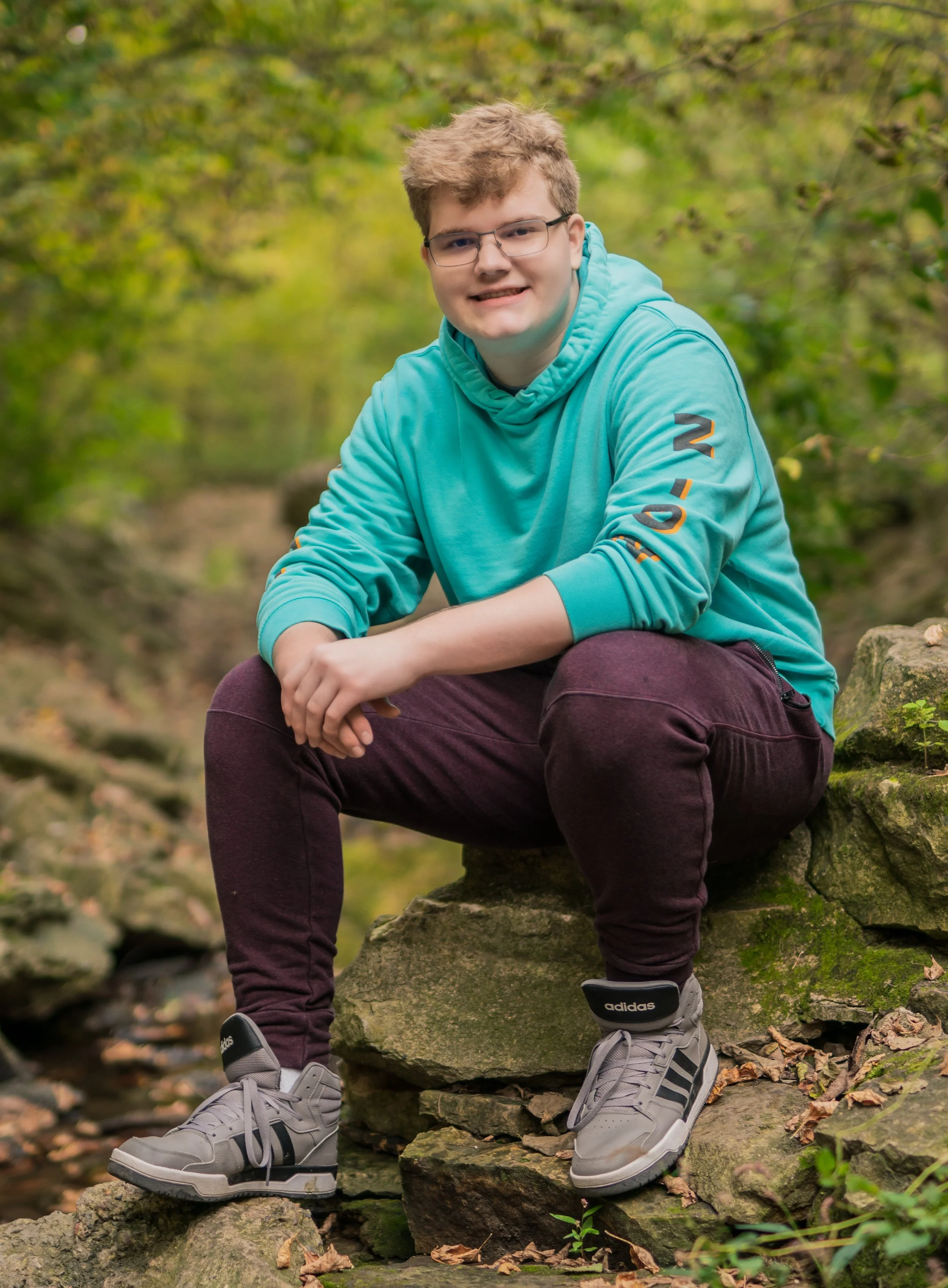 A young man with glasses and blond hair smiling while sitting on rocks in a wooded outdoor area.