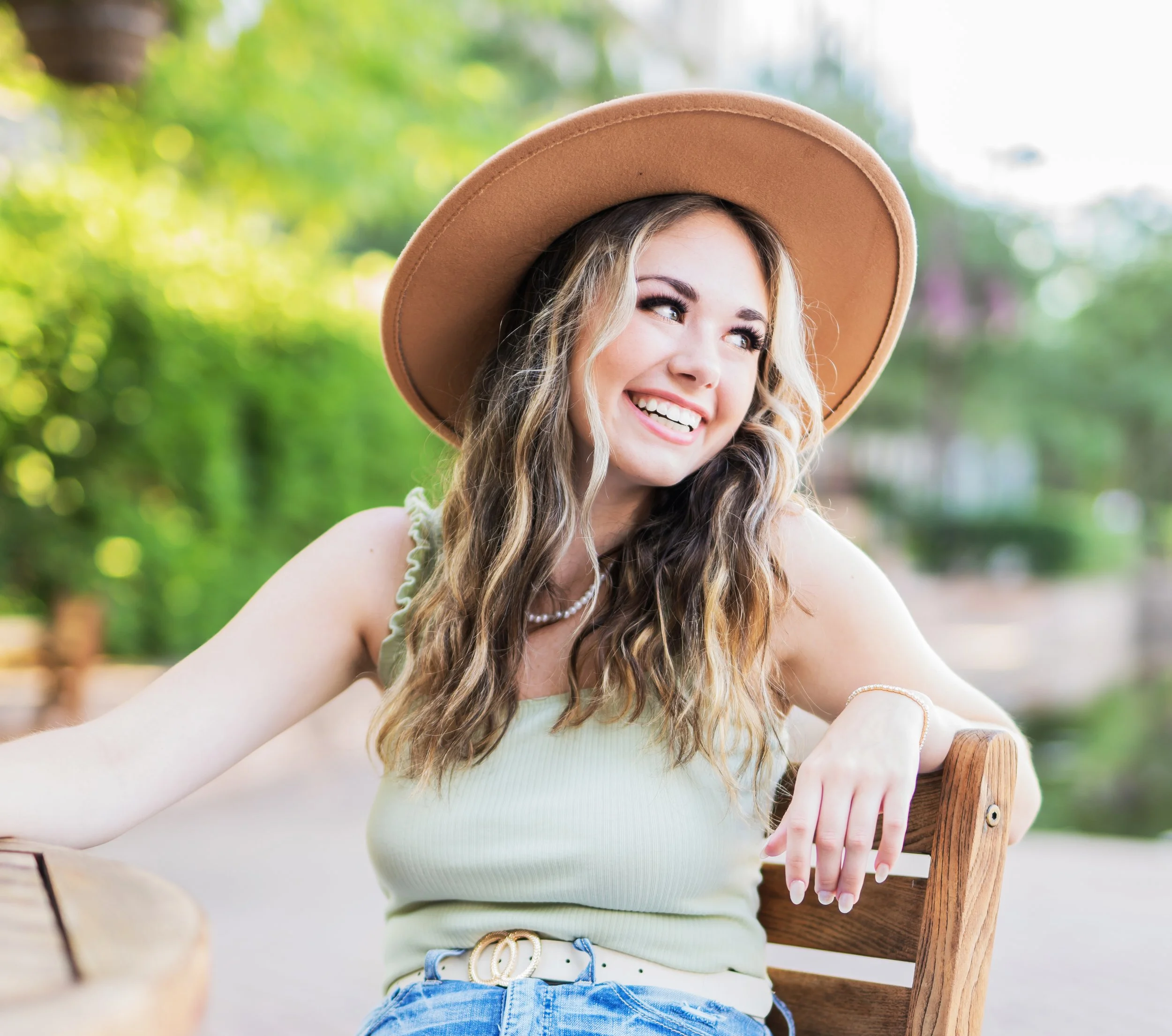 A smiling young woman with wavy brown hair wearing a wide-brimmed tan hat, sitting outdoors on a wooden bench with a blurred green and yellow background.