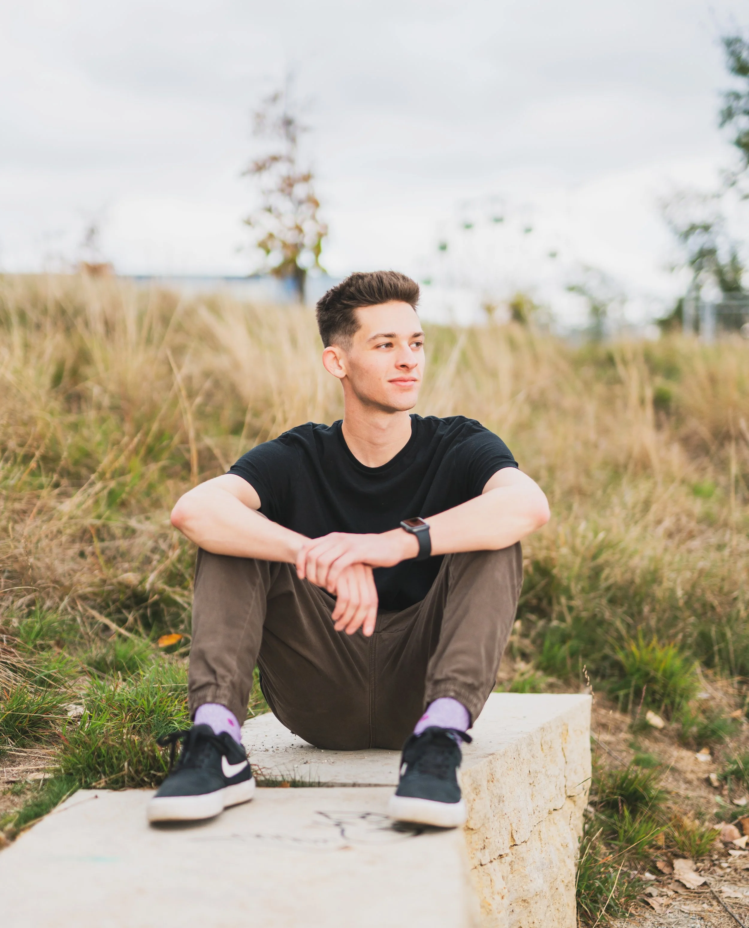 Young man sitting cross-legged on a concrete block outdoors, wearing a black t-shirt, brown pants, black sneakers, and a watch, with tall grass and trees in the background.