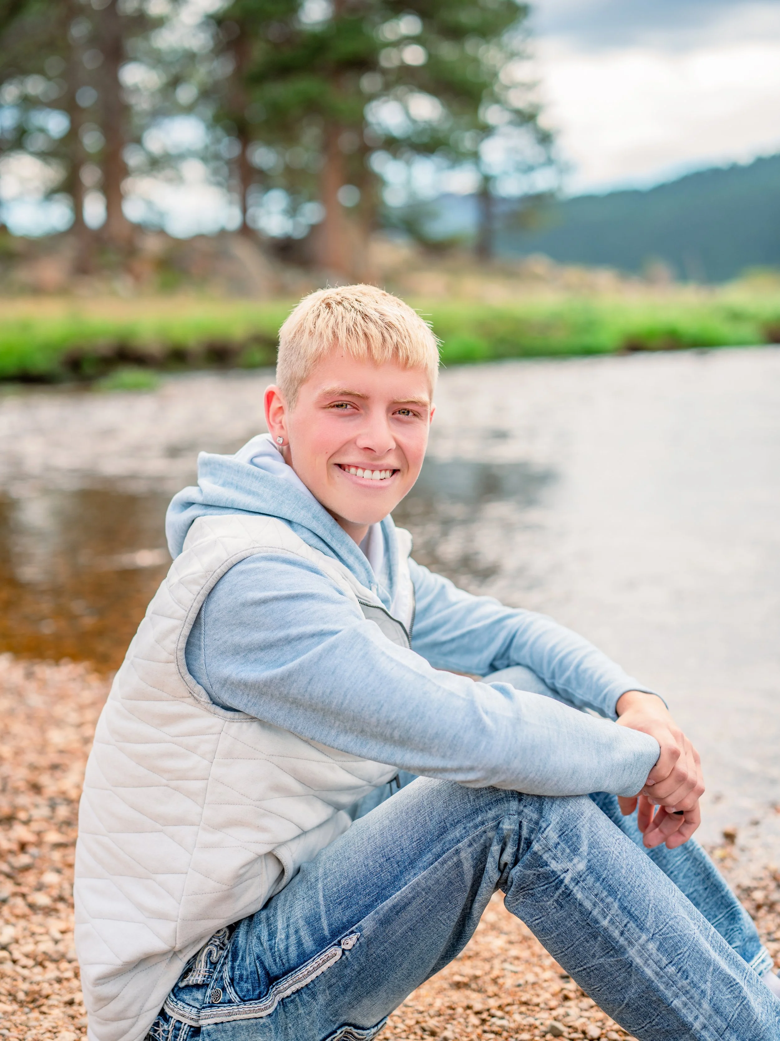 A smiling young man with short blonde hair sitting near a riverbank with trees in the background.