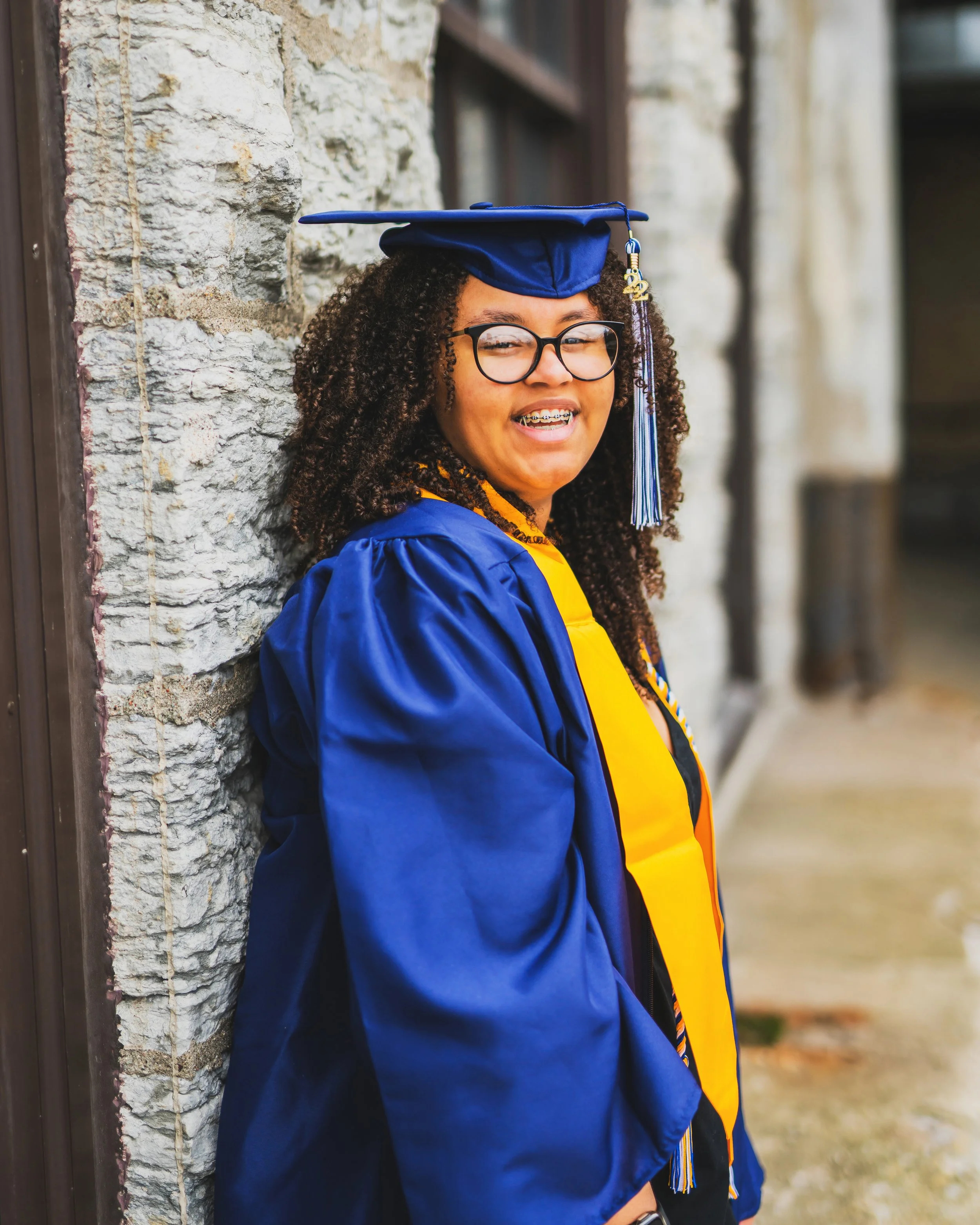 Young woman in graduation cap and gown smiling, leaning against a stone wall outdoors.