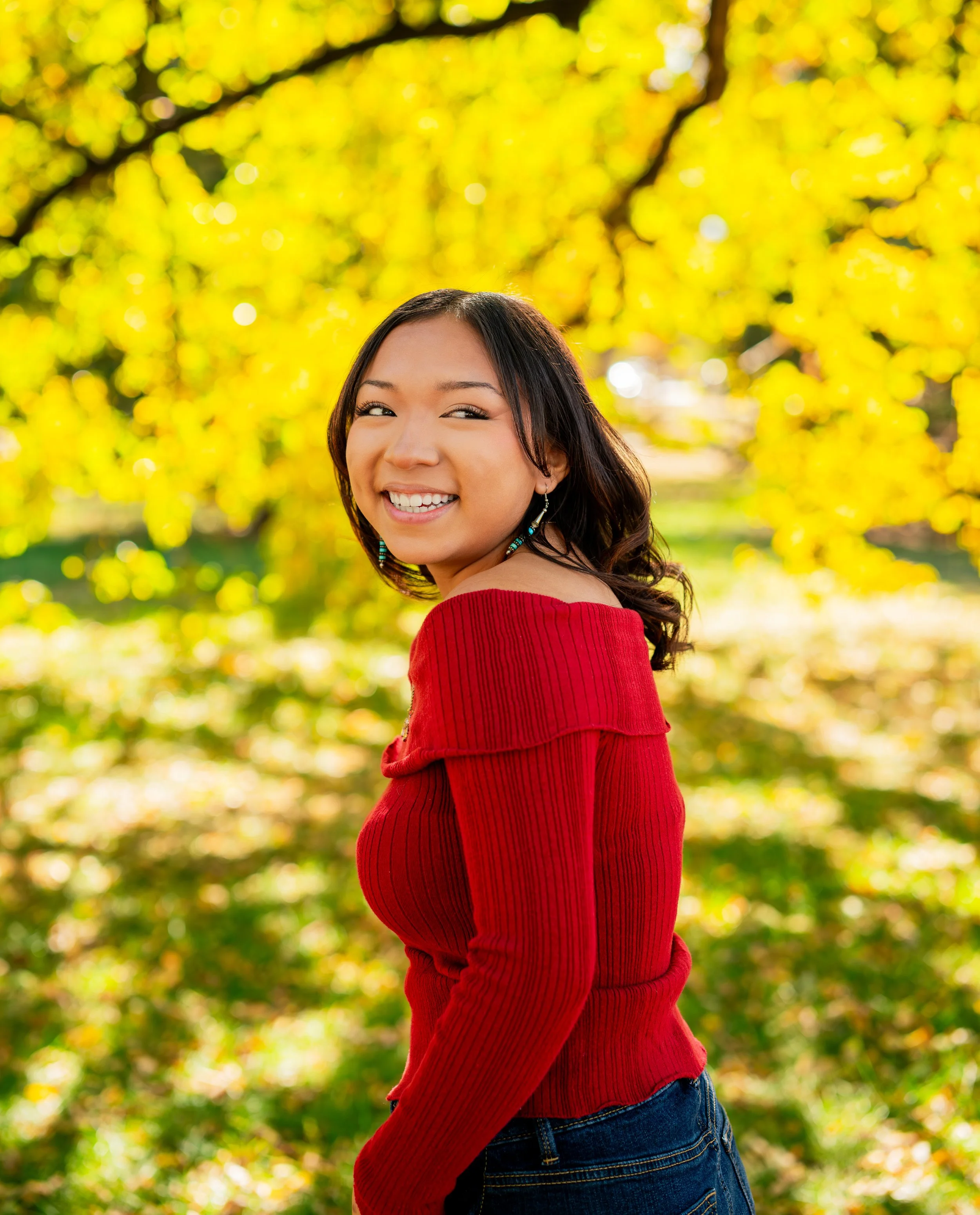 A young woman with medium-length dark hair, wearing a red sweater and blue jeans, smiling outdoors with yellow autumn leaves in the background.