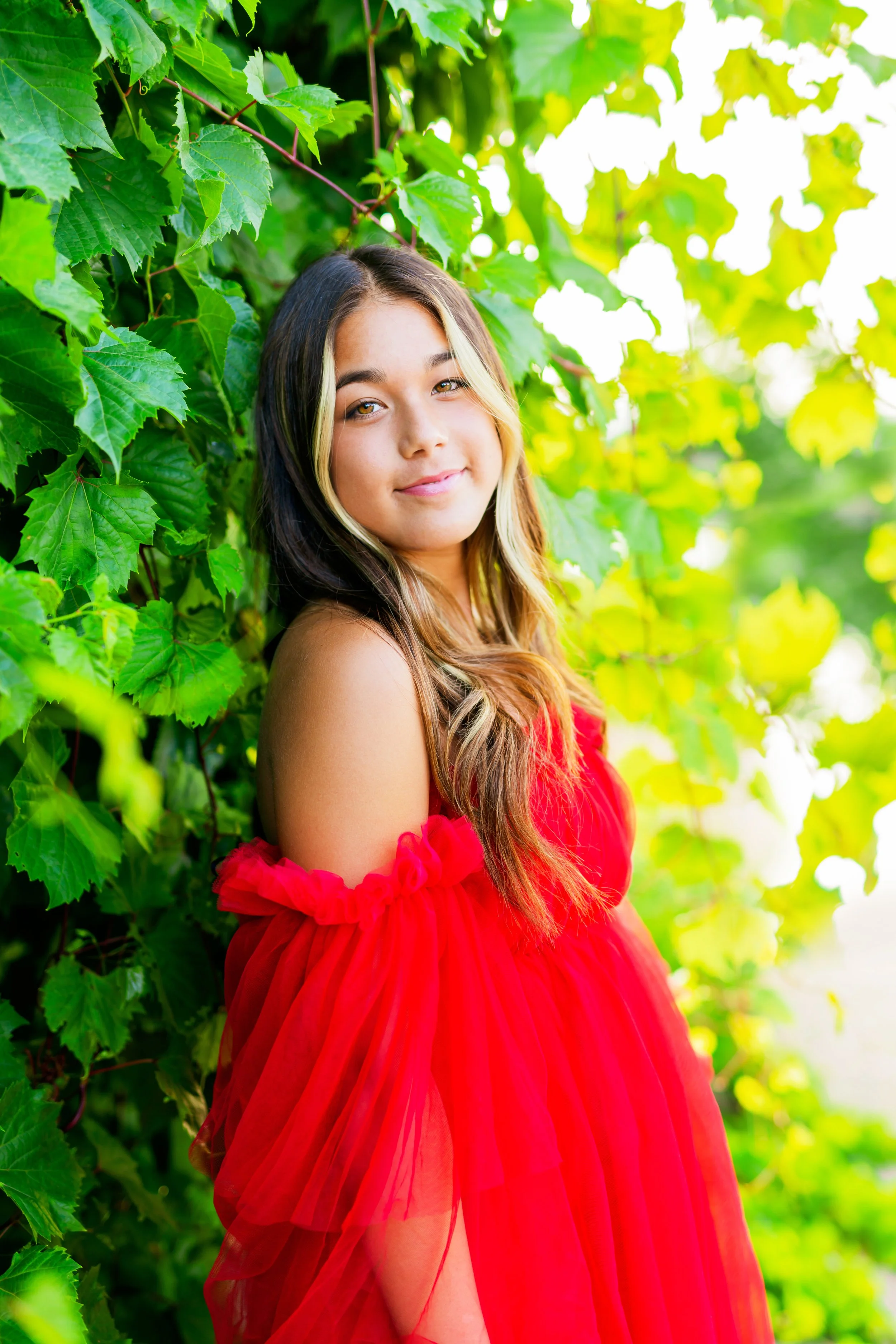 A young girl with long, wavy hair wearing a red dress with off-shoulder ruffles, standing next to lush green leafy vines, smiling softly at the camera.