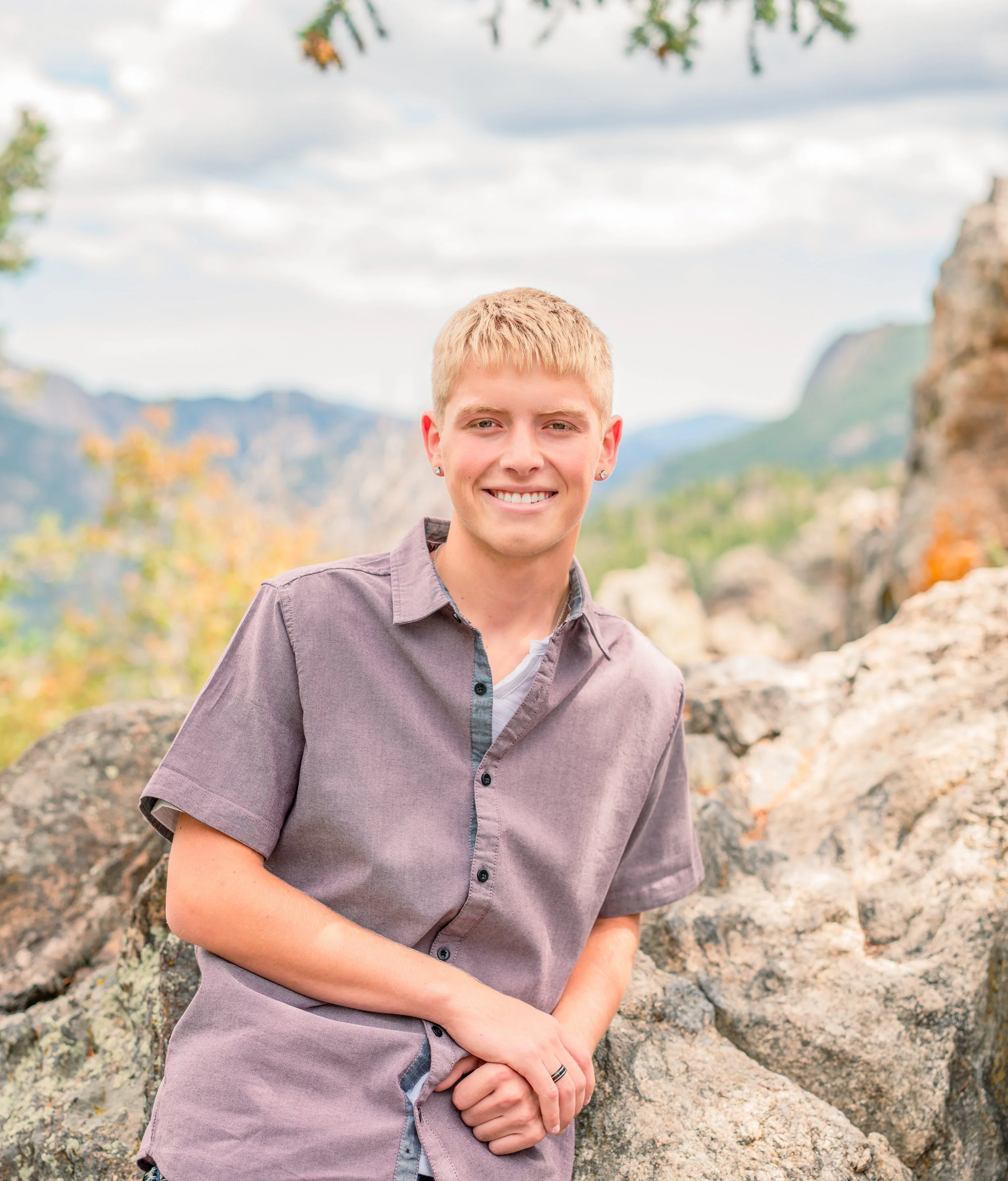 A young man with blonde hair smiling outdoors, standing near rocks with a mountain and cloudy sky in the background.