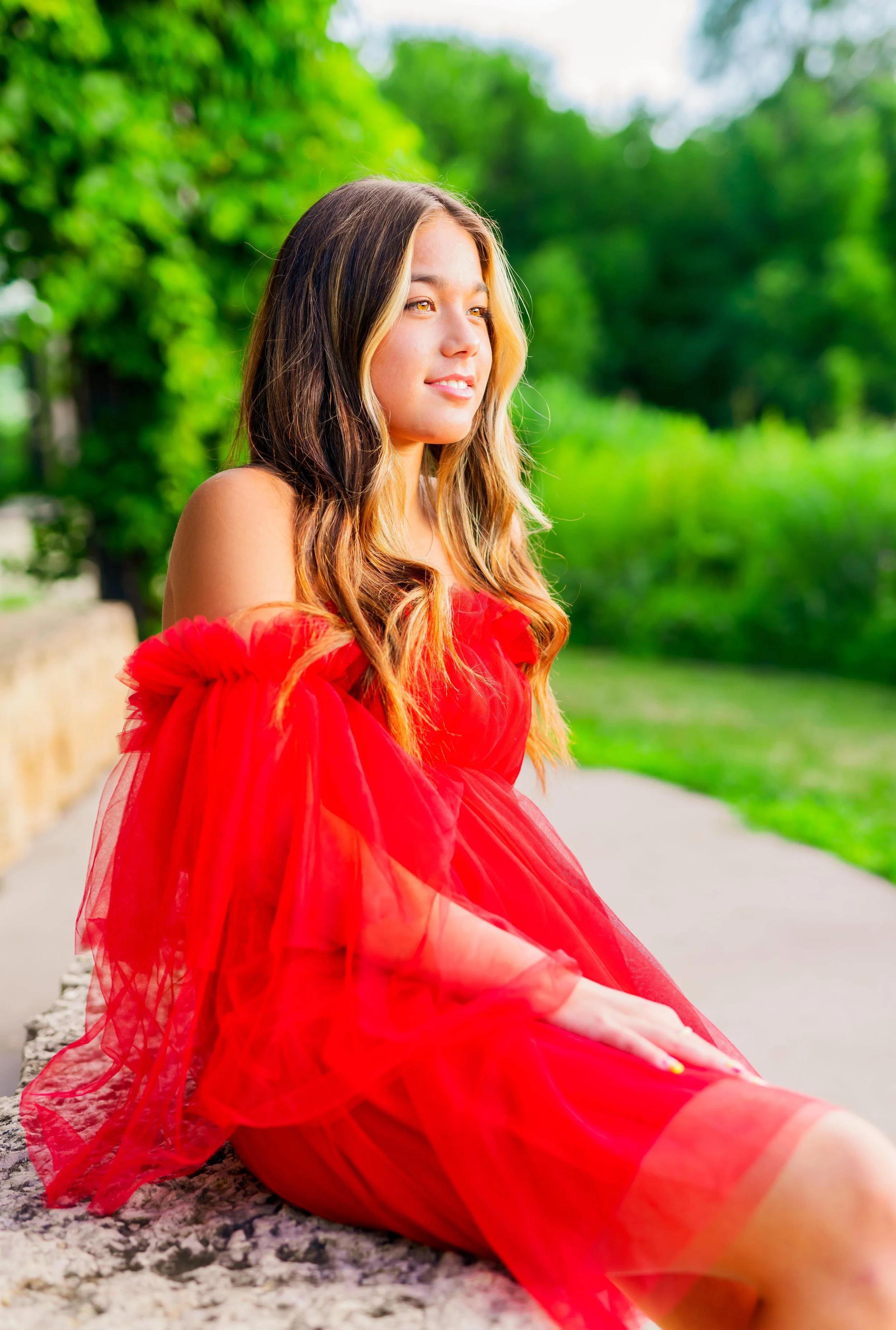 Young woman wearing a red dress sitting outdoors on a stone ledge with green trees in the background.