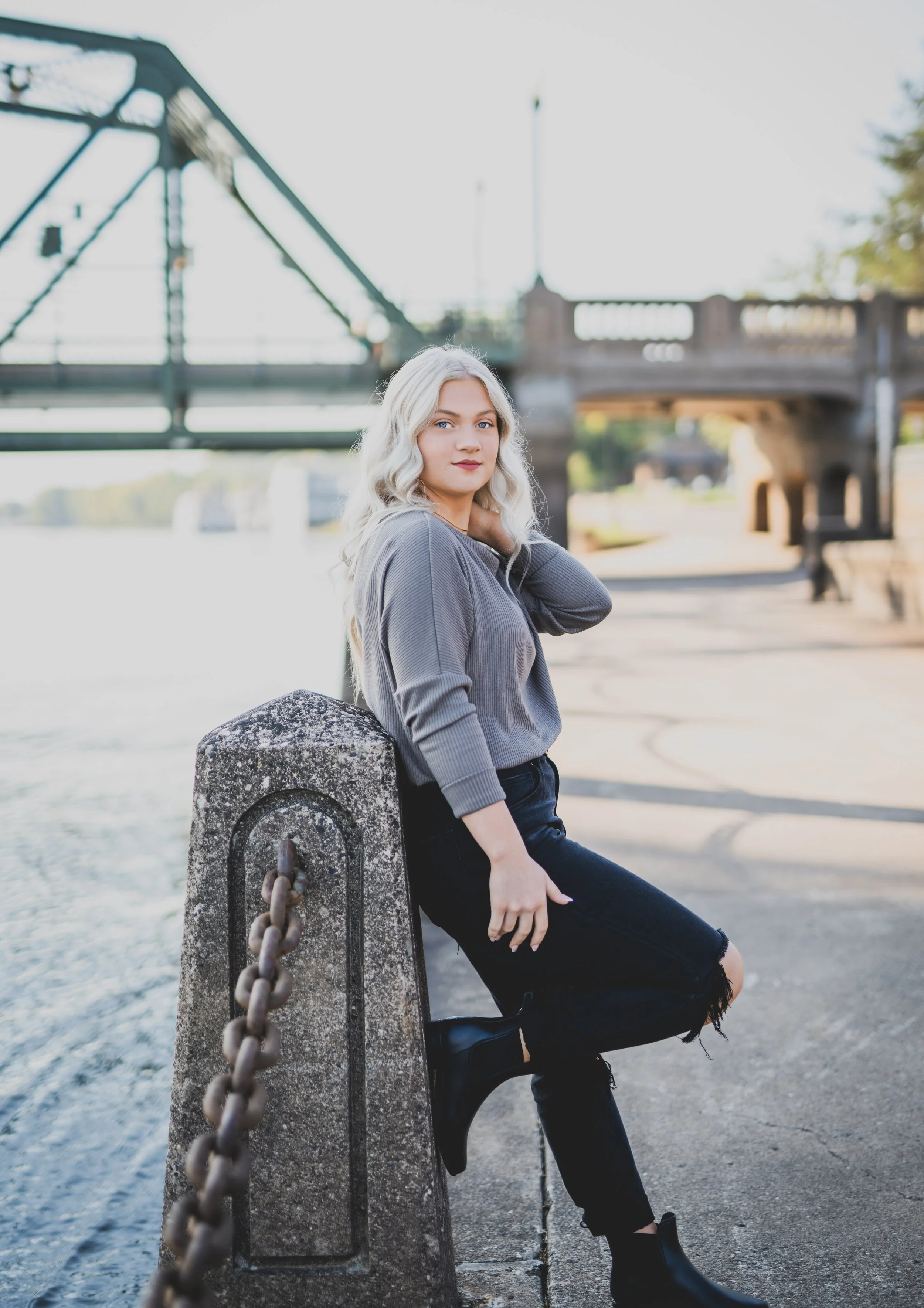 Young woman with platinum blonde hair leaning against a concrete bollard with a metal chain near a river, with a bridge in the background.