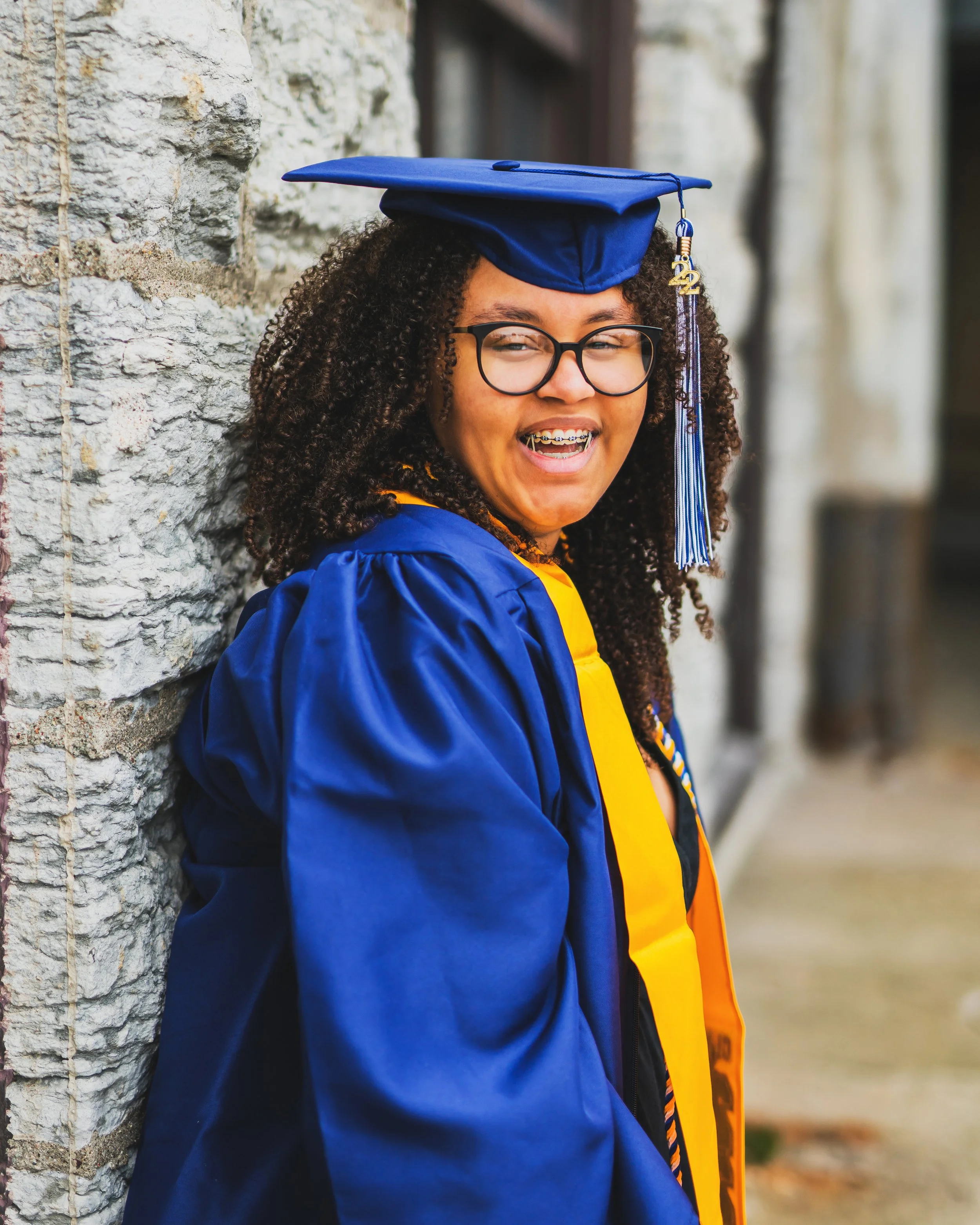 A young woman in cap and gown leaning against a stone wall, smiling, wearing glasses and braces.