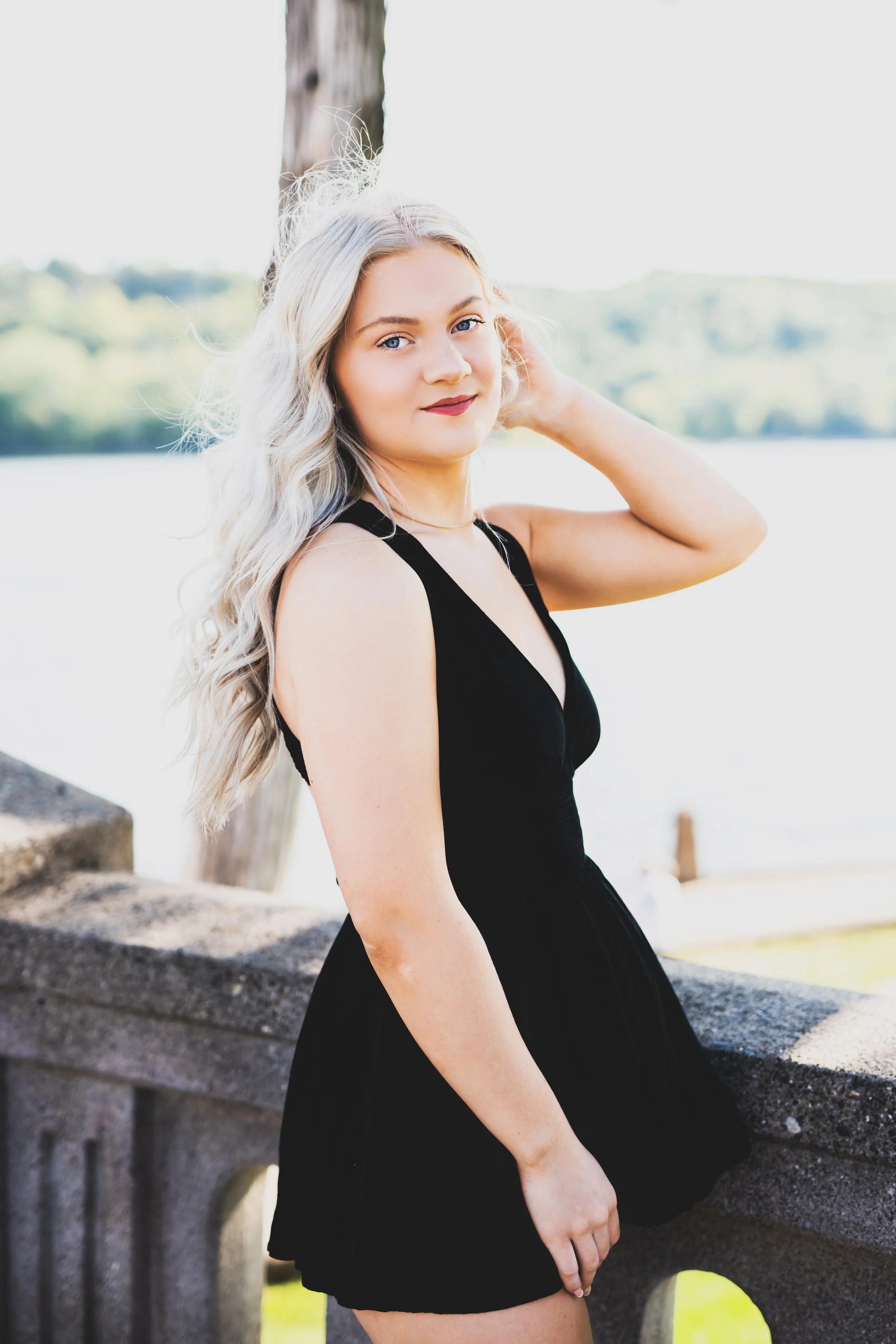 A young woman in a black dress standing outdoors near water, with trees in the background, posing by a concrete railing.