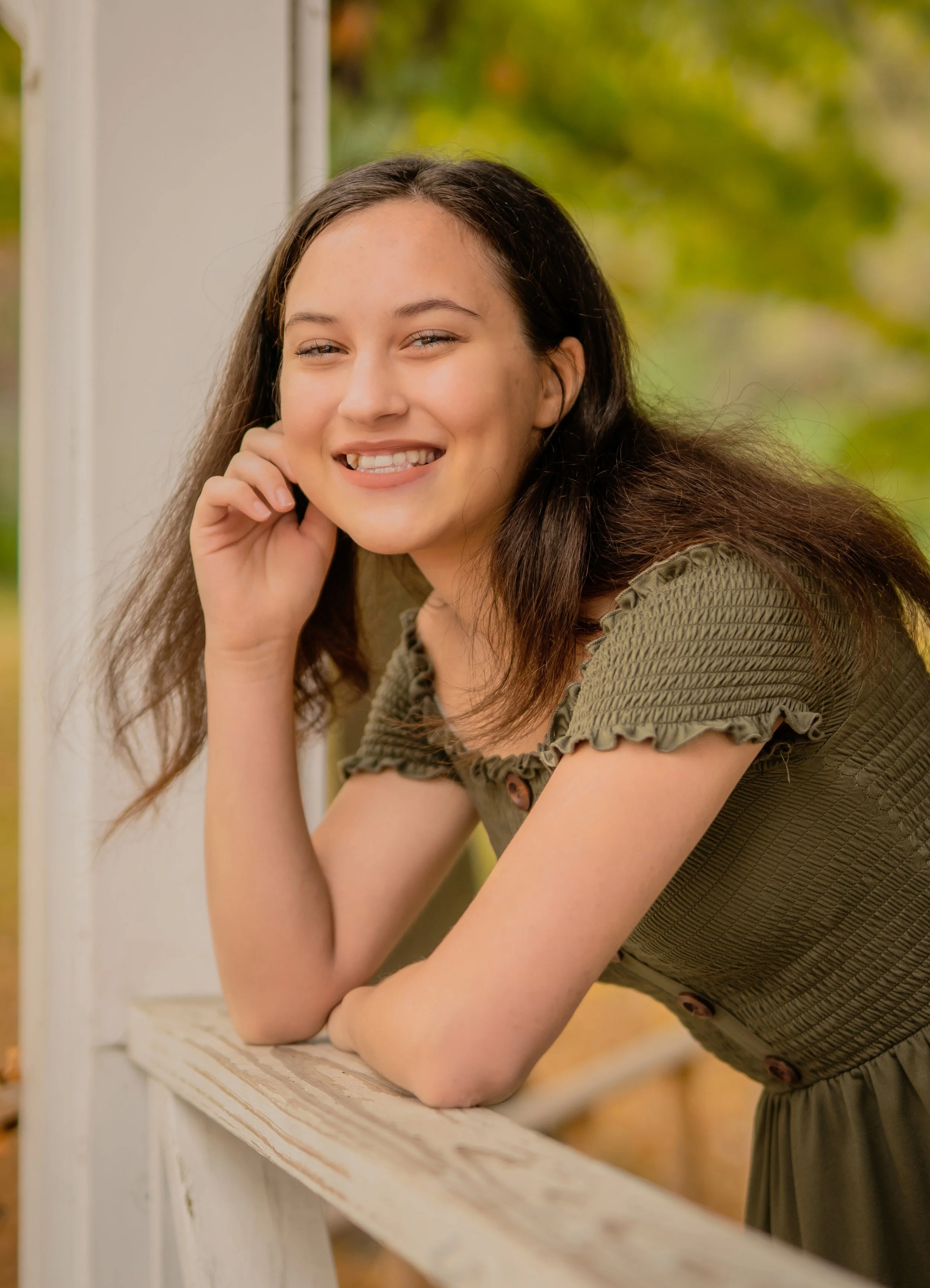 A smiling young woman with long brown hair leaning on a white railing outdoors during fall, with trees and yellow leaves in the background.