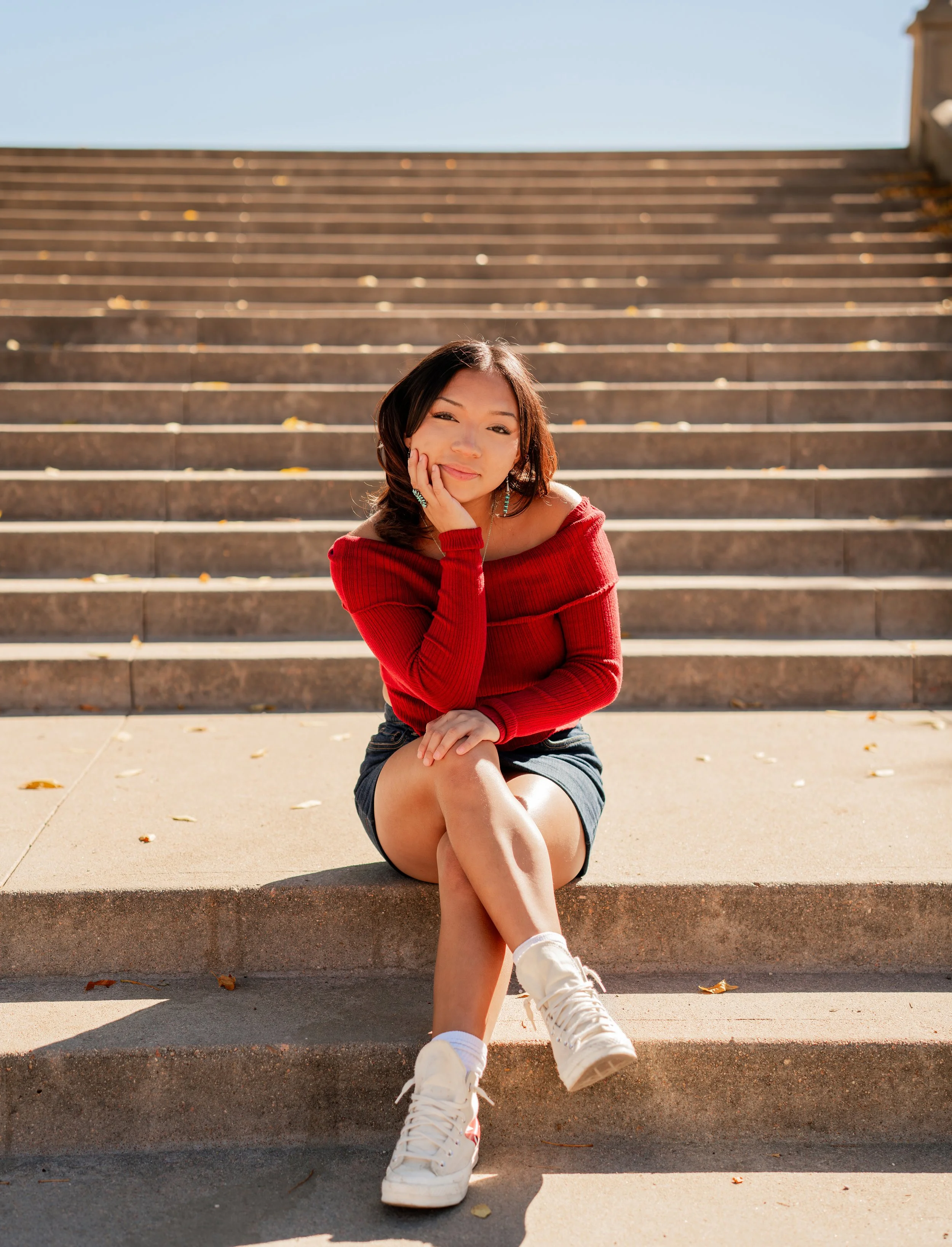 A young woman with shoulder-length dark hair sitting on outdoor steps, wearing a red off-shoulder sweater, black shorts, and white sneakers, smiling and resting her chin on her hand.
