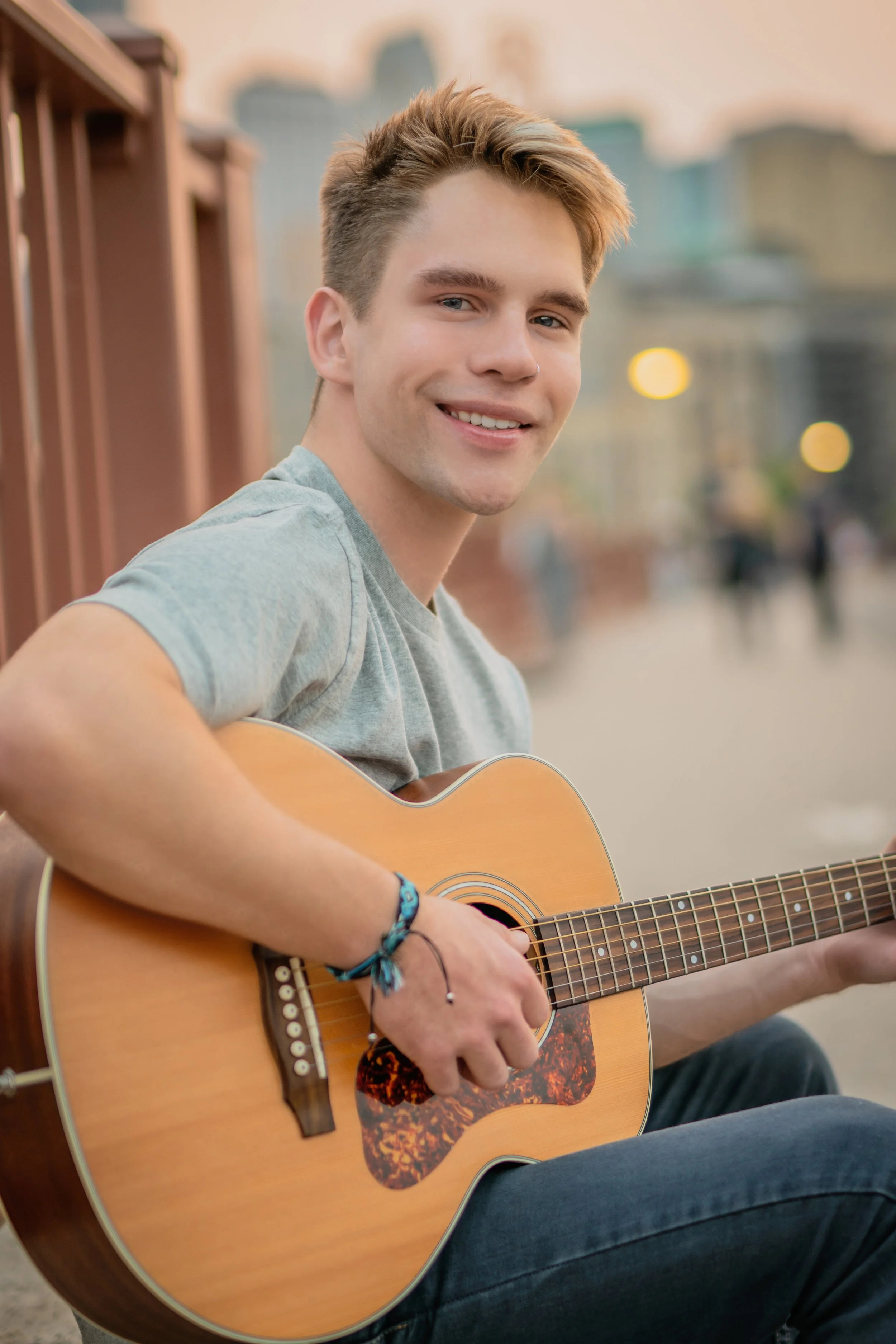 A young man smiling and playing an acoustic guitar outdoors in an urban setting during sunset.
