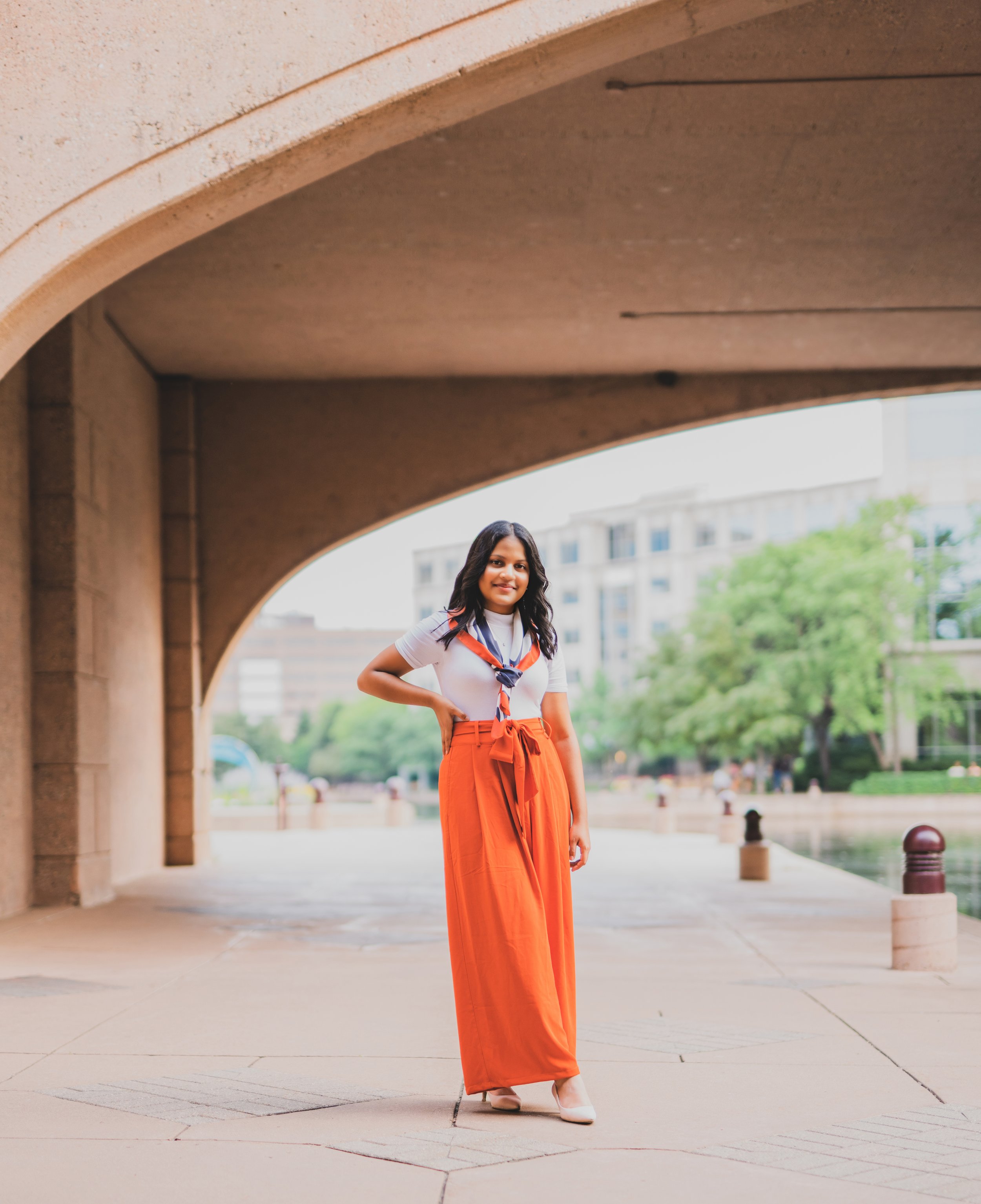 A woman standing under a concrete archway near a body of water with greenery and buildings in the background.