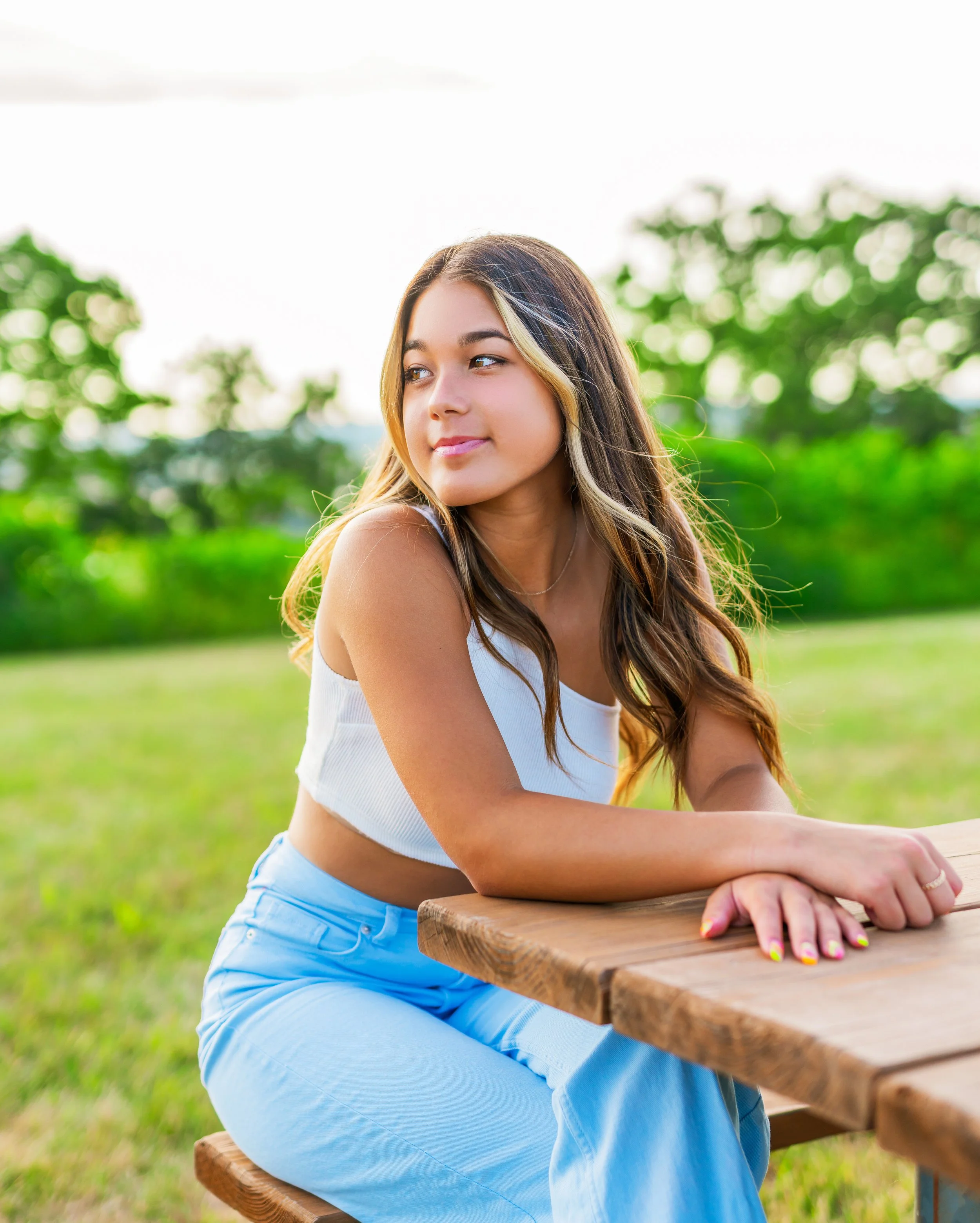 A young woman with long, wavy brown hair and light skin sitting at a wooden picnic table, outdoors in a green park, looking to the side with a slight smile.