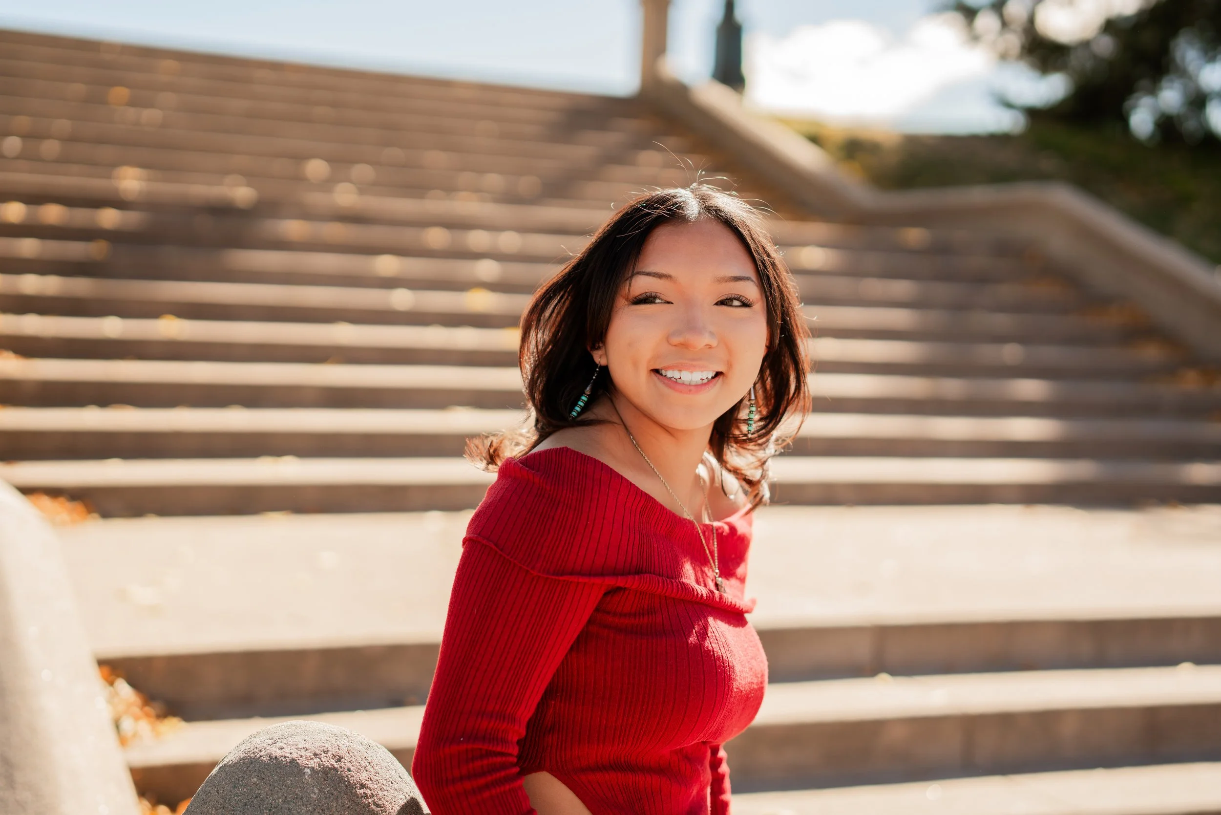 A young woman with shoulder-length dark hair smiling while standing outdoors in front of a staircase, wearing a red off-shoulder top, earrings, and a necklace.