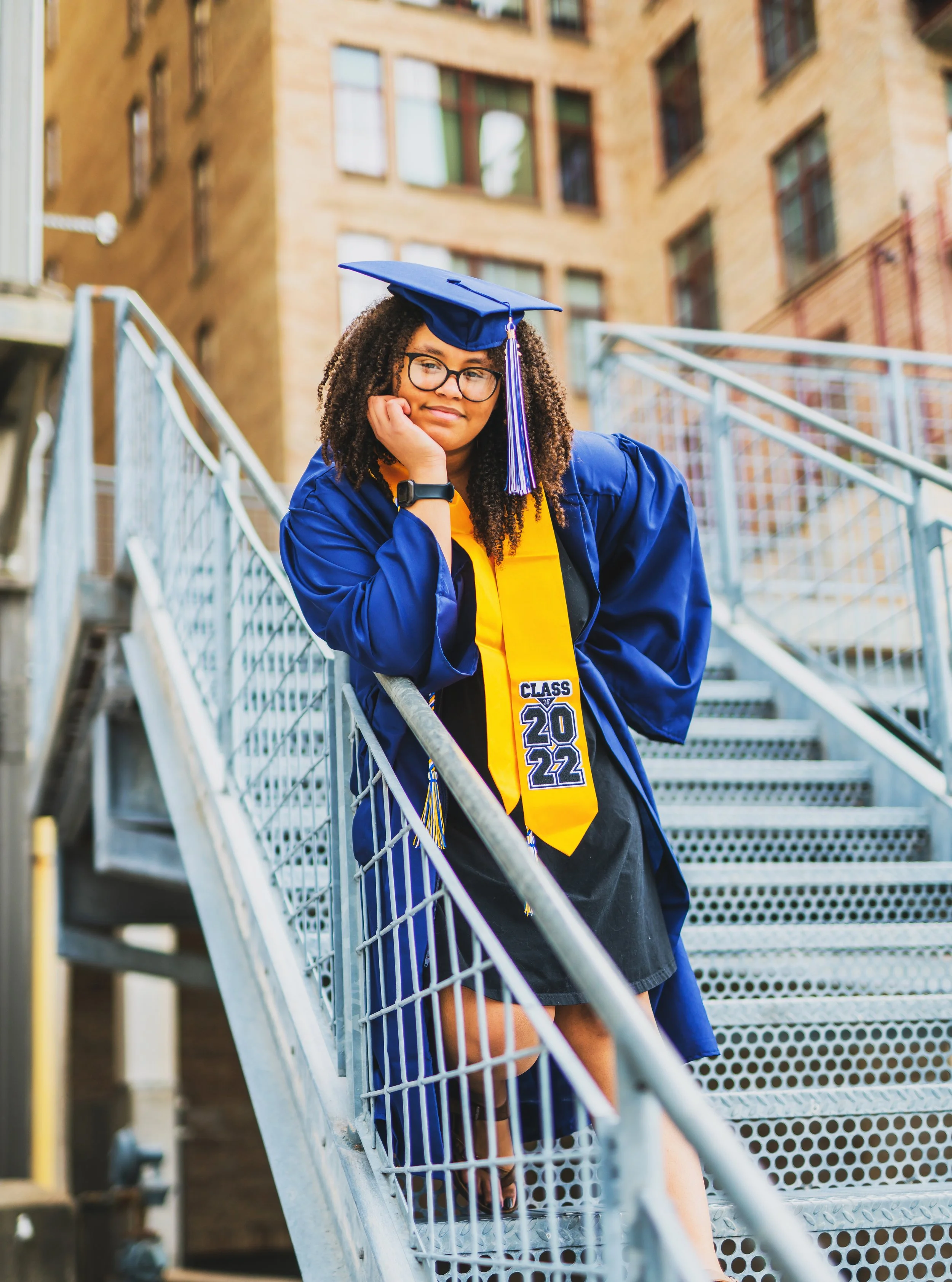 A young woman in graduation cap and gown, wearing a yellow stole that says 'Class of 2022,' standing on outdoor metal stairs with her hand resting on her face, smiling softly.