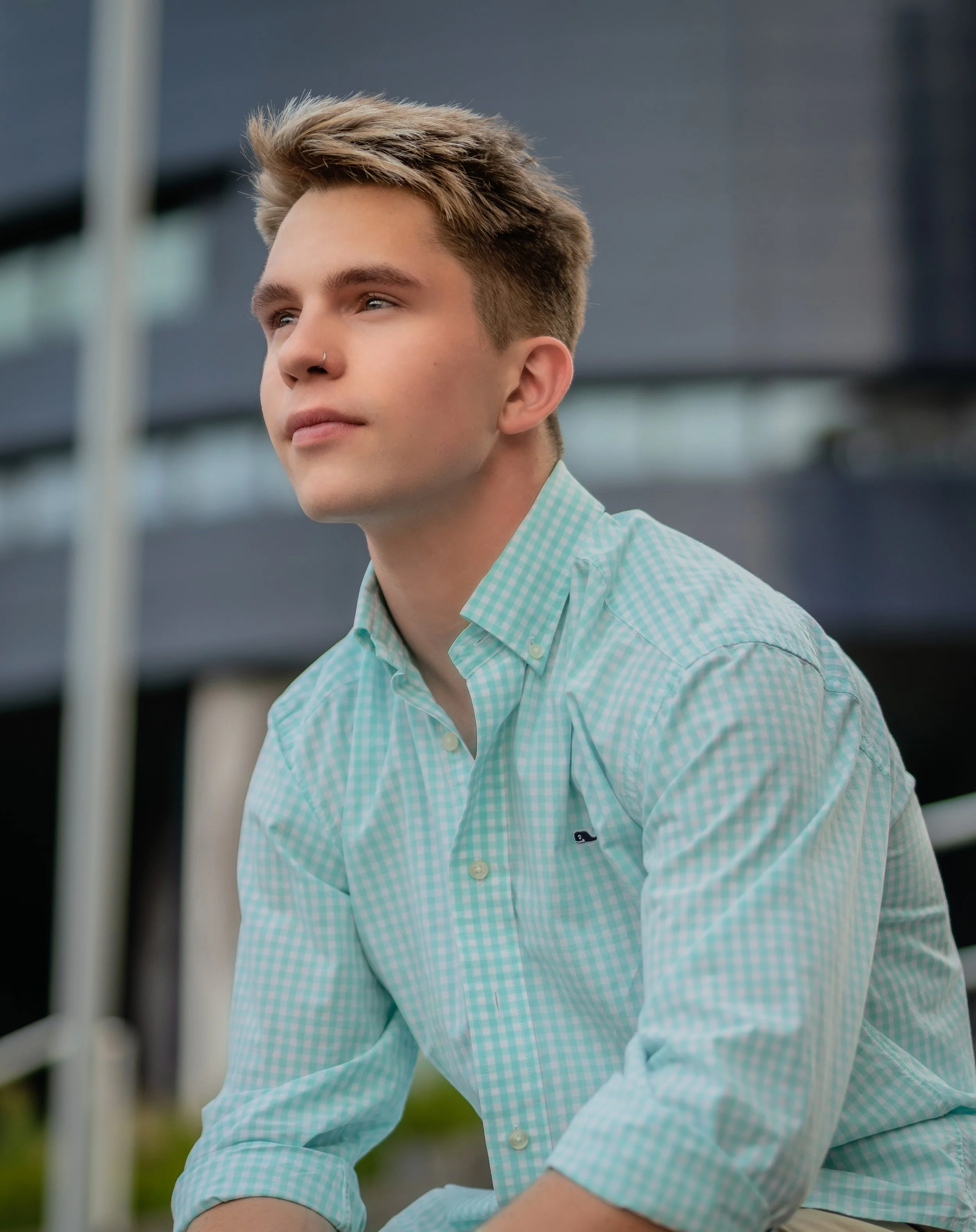 A young man with blond hair wearing a light green checkered shirt, sitting outdoors with modern buildings in the background.