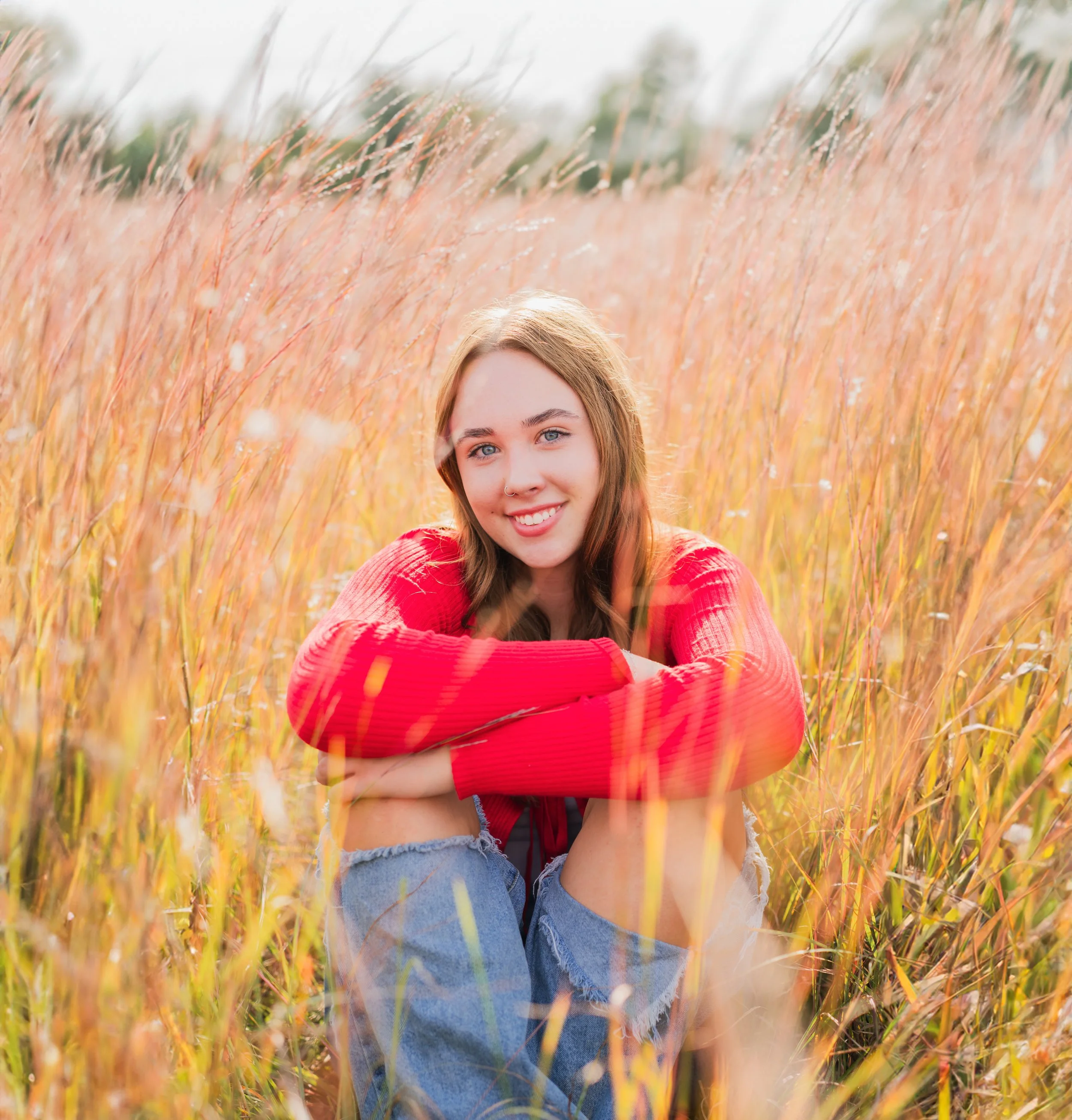 A young woman with brown hair, wearing a red sweater, sitting in a tall, golden grass field, smiling at the camera.