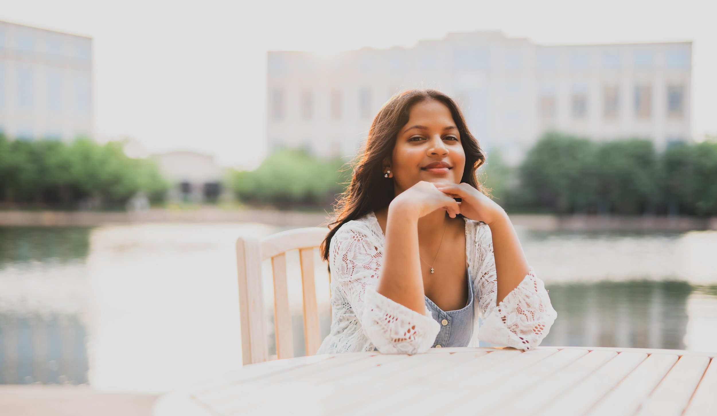Young woman sitting at an outdoor table near a river, smiling at the camera with city buildings in the background during daytime.