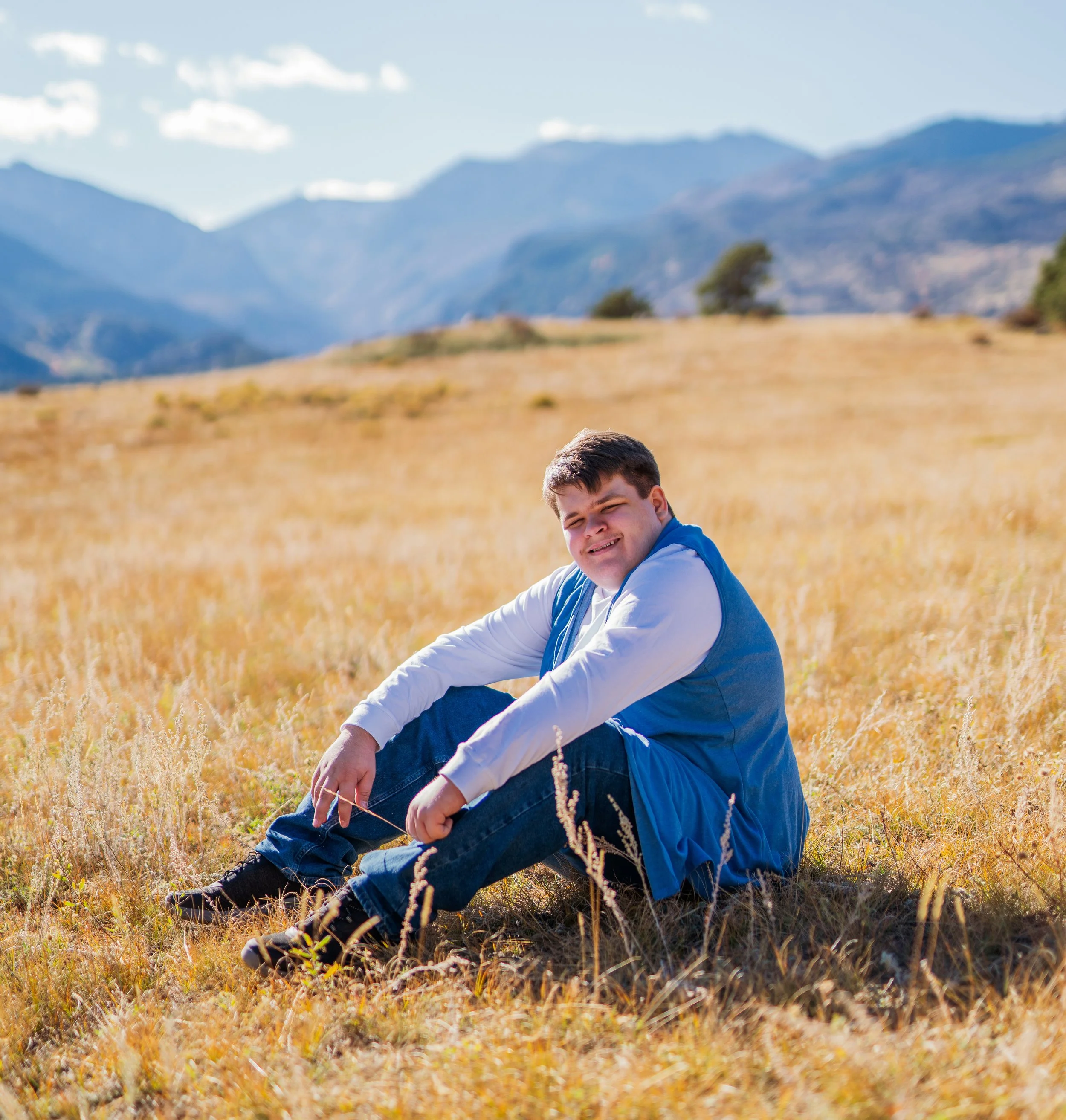A young man sitting in a grassy field with mountains in the background.