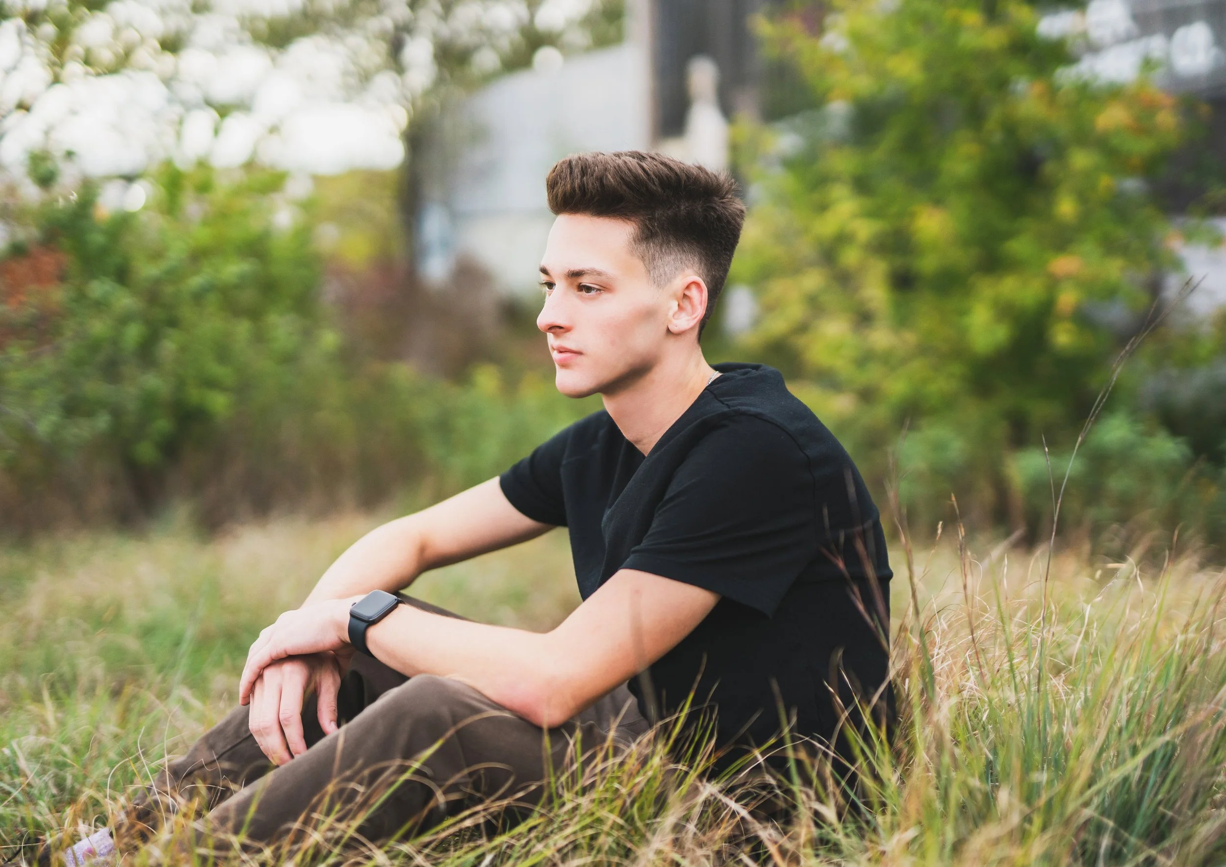 A young man sitting in a grassy field with trees in the background, wearing a black t-shirt and a smartwatch.