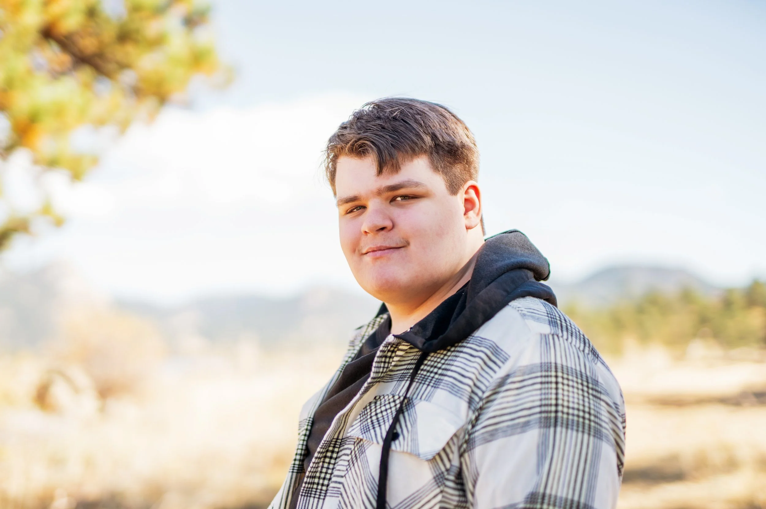 Young man outdoors with a blurred natural background, wearing a plaid jacket and a hoodie, smiling slightly.