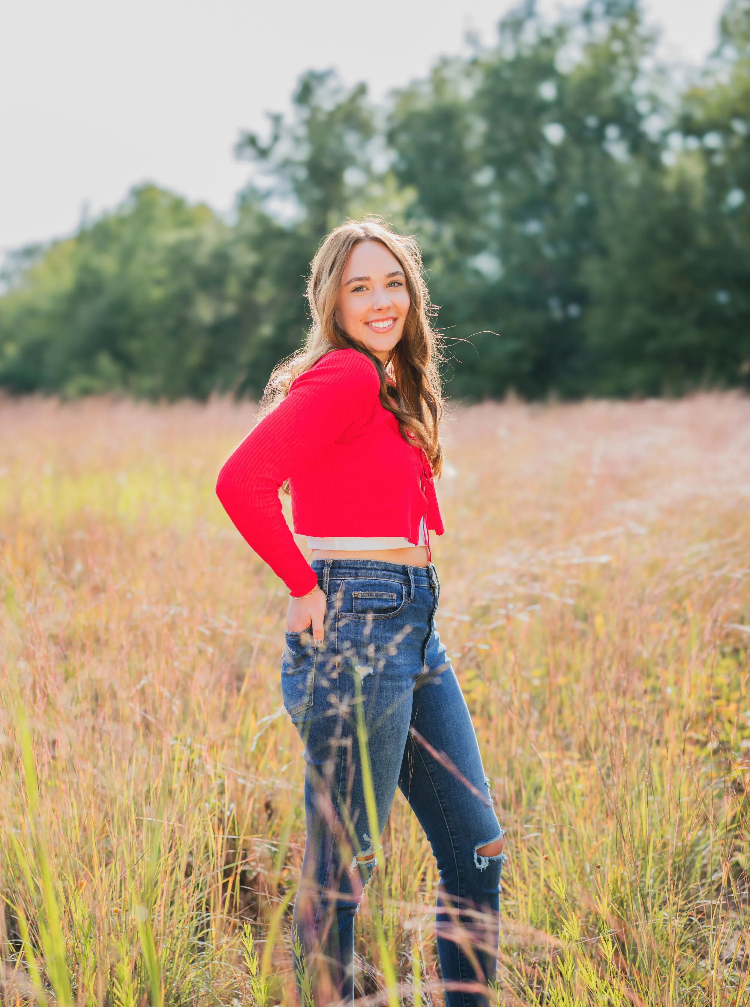 A young woman in a red cropped sweater and ripped jeans standing in a field of tall grass, smiling at the camera with trees in the background on a sunny day.