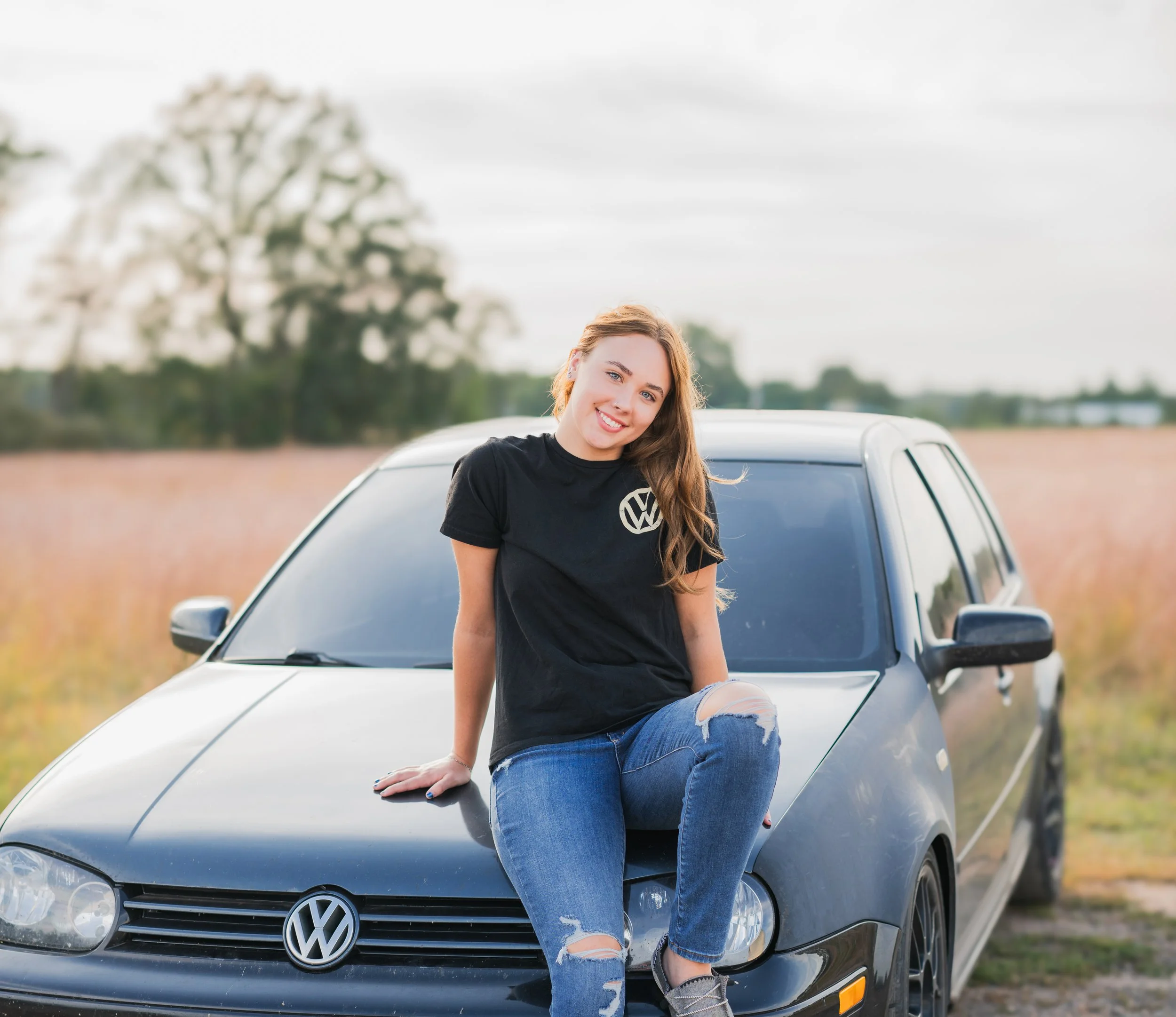 Young woman with long hair sits on the hood of a black Volkswagen car in a field with trees in the background.