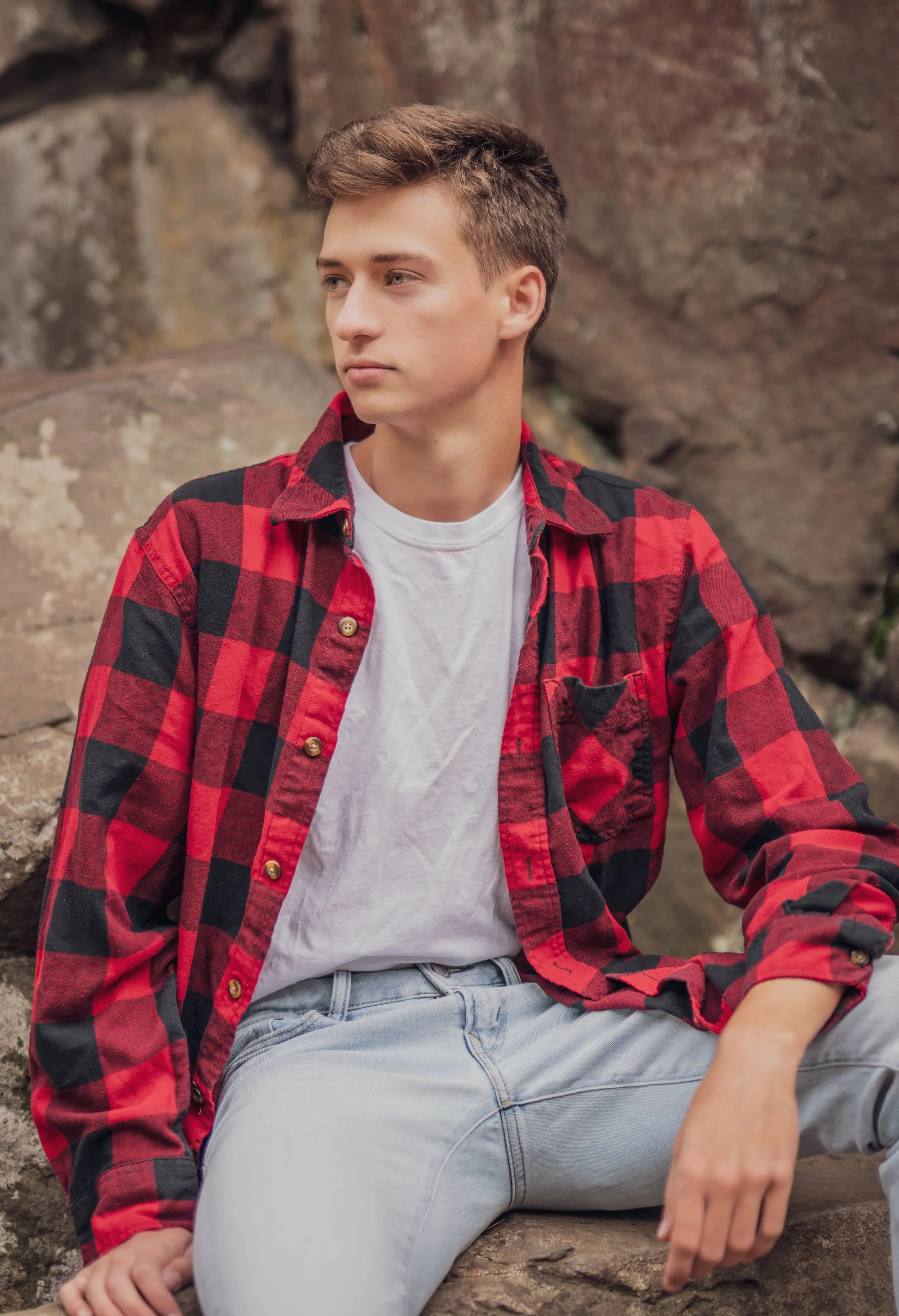 A young man sitting on a large rock outdoors, wearing a red and black checkered flannel shirt over a white T-shirt and light-wash jeans, looking off to the side with a thoughtful expression.