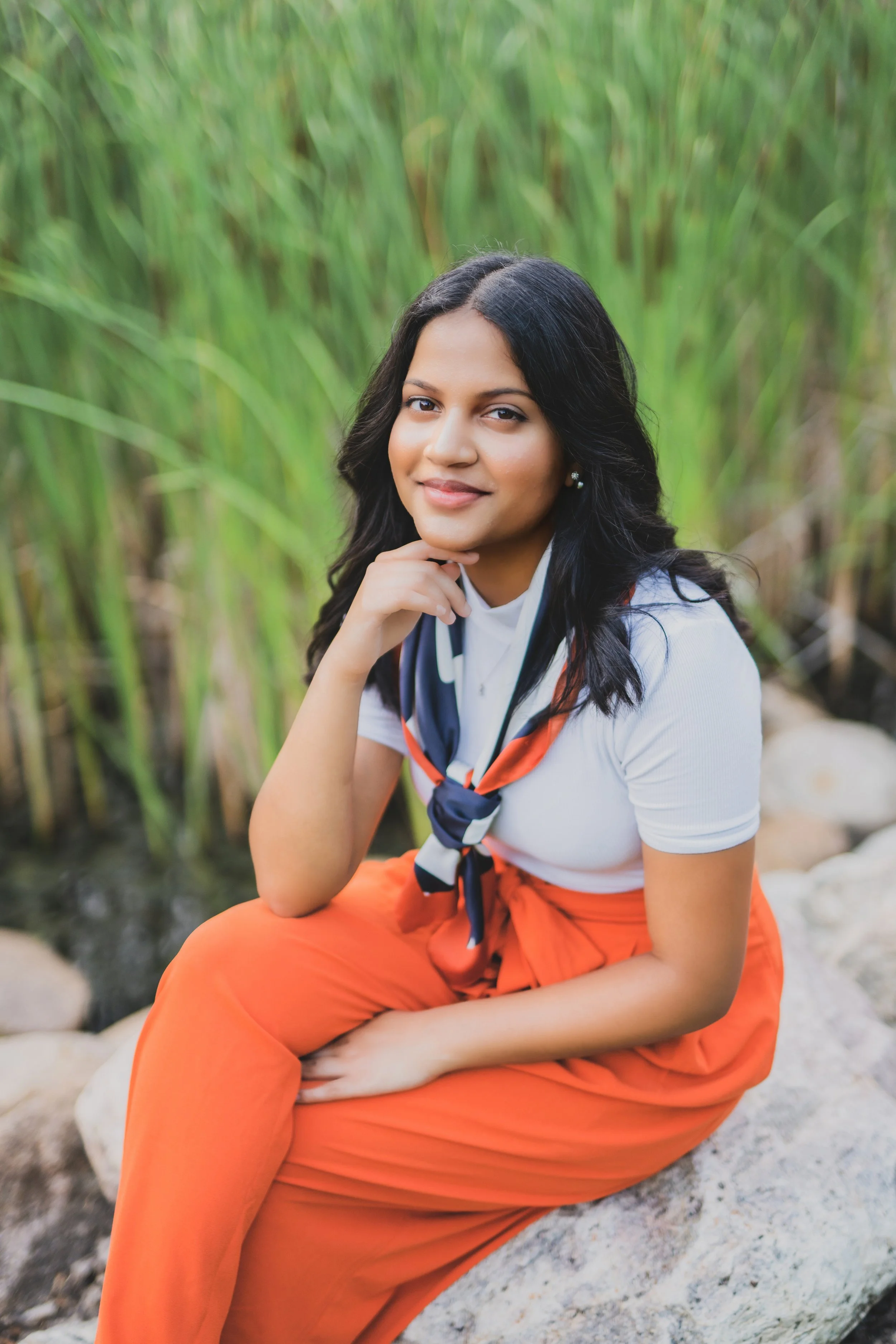 A young woman sitting on a large rock outdoors, surrounded by tall green grass, wearing an orange skirt, white top, and a blue and orange neckerchief, smiling at the camera.
