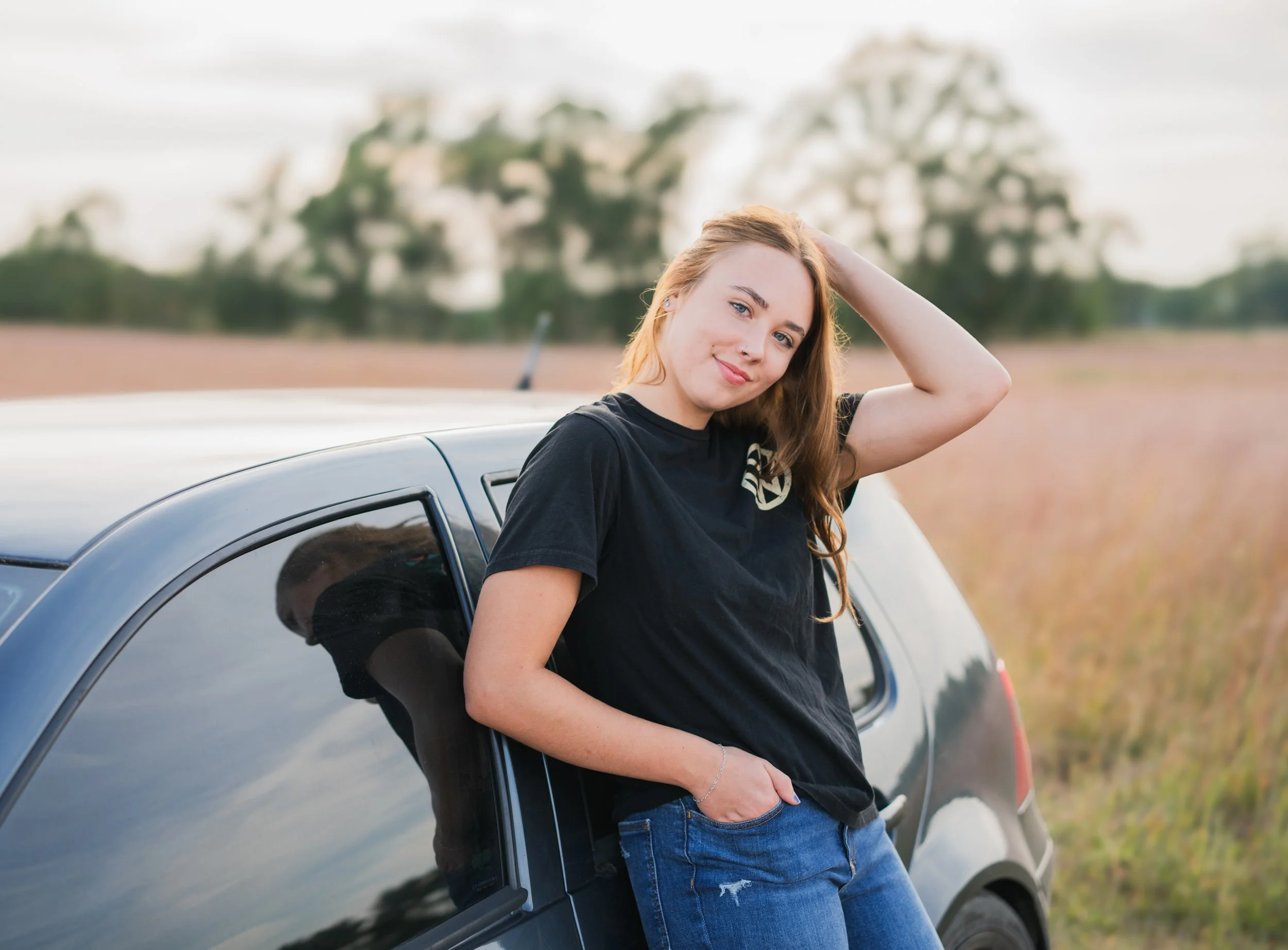 A young woman with long light brown hair, wearing a black t-shirt and blue jeans, leaning against a black car in an open field during daytime, with a slight smile and her hand resting on her head.