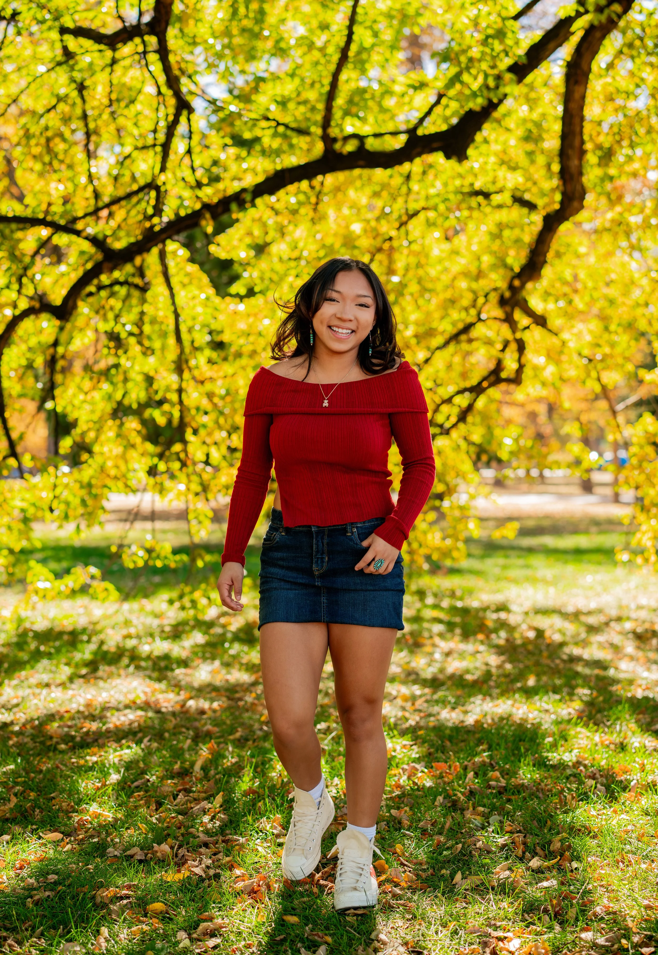 A young woman smiling and walking outdoors in a park with autumn leaves on the ground and a large tree with yellow leaves in the background. She is wearing a red off-the-shoulder sweater, denim skirt, white sneakers, and jewelry.