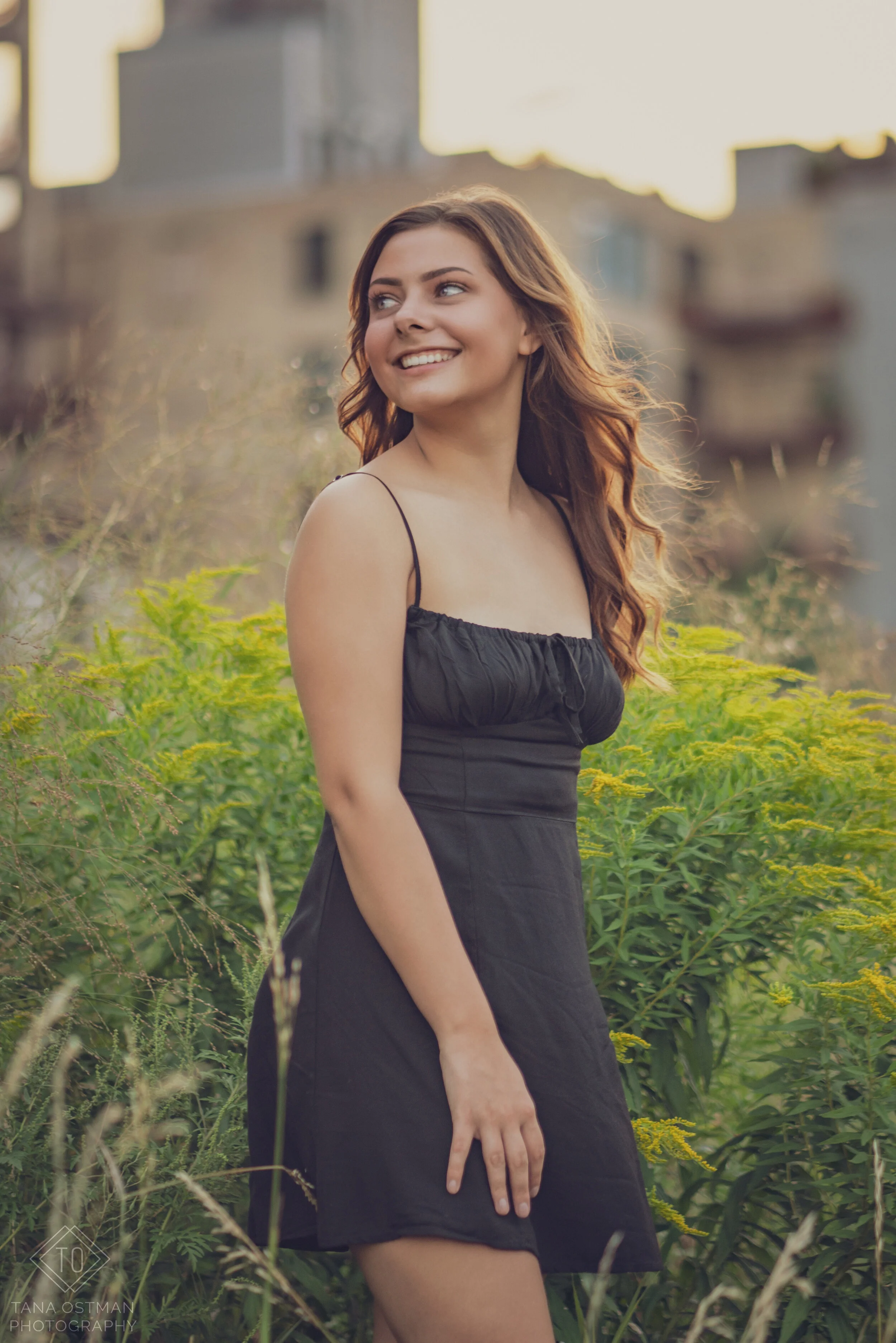 Young woman in a black dress smiling in a green outdoor setting during sunset.