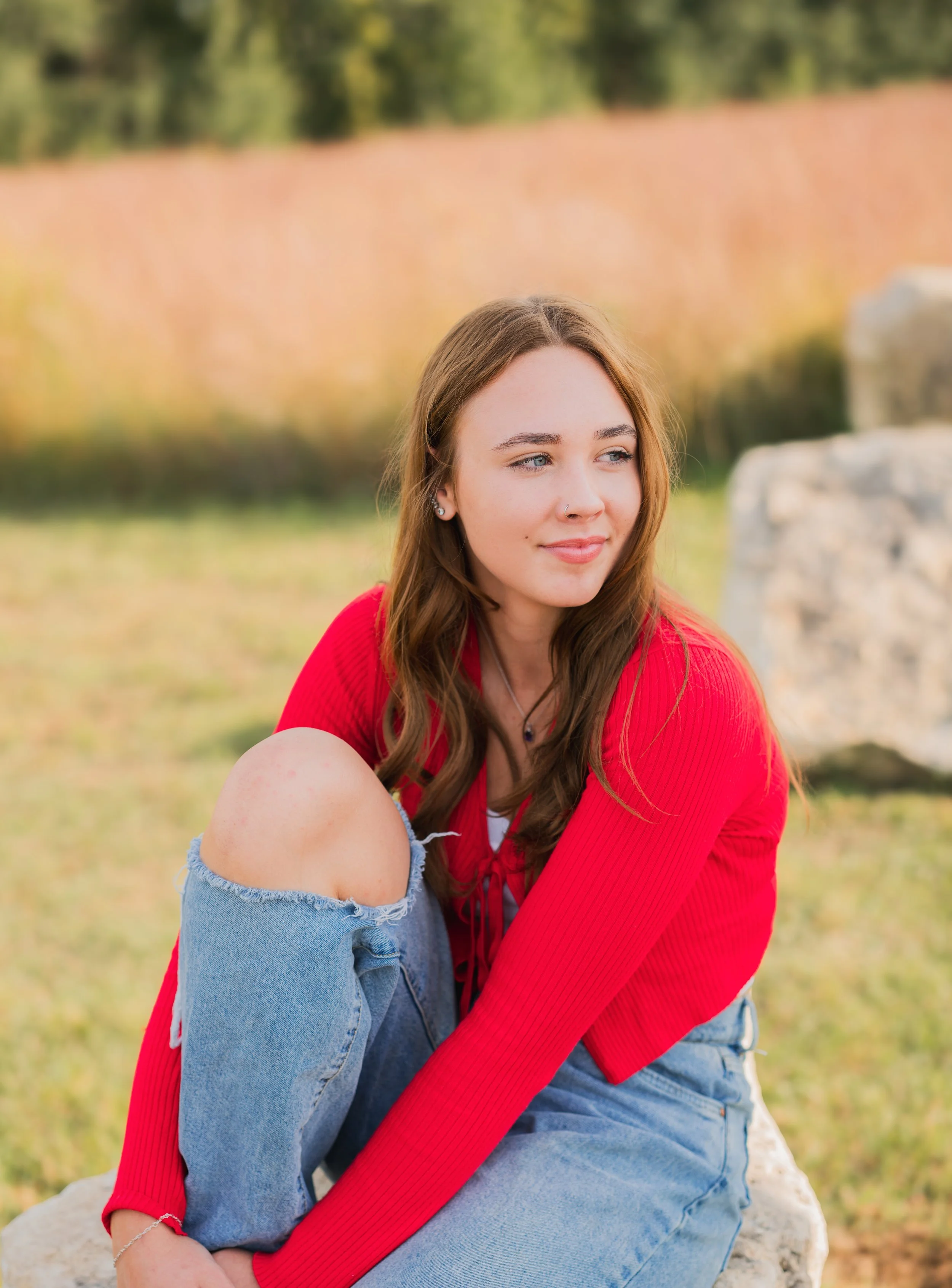 A young woman with long brown hair, wearing a red cardigan, ripped jeans, and a necklace, sitting outdoors on a rock with a blurred natural background.