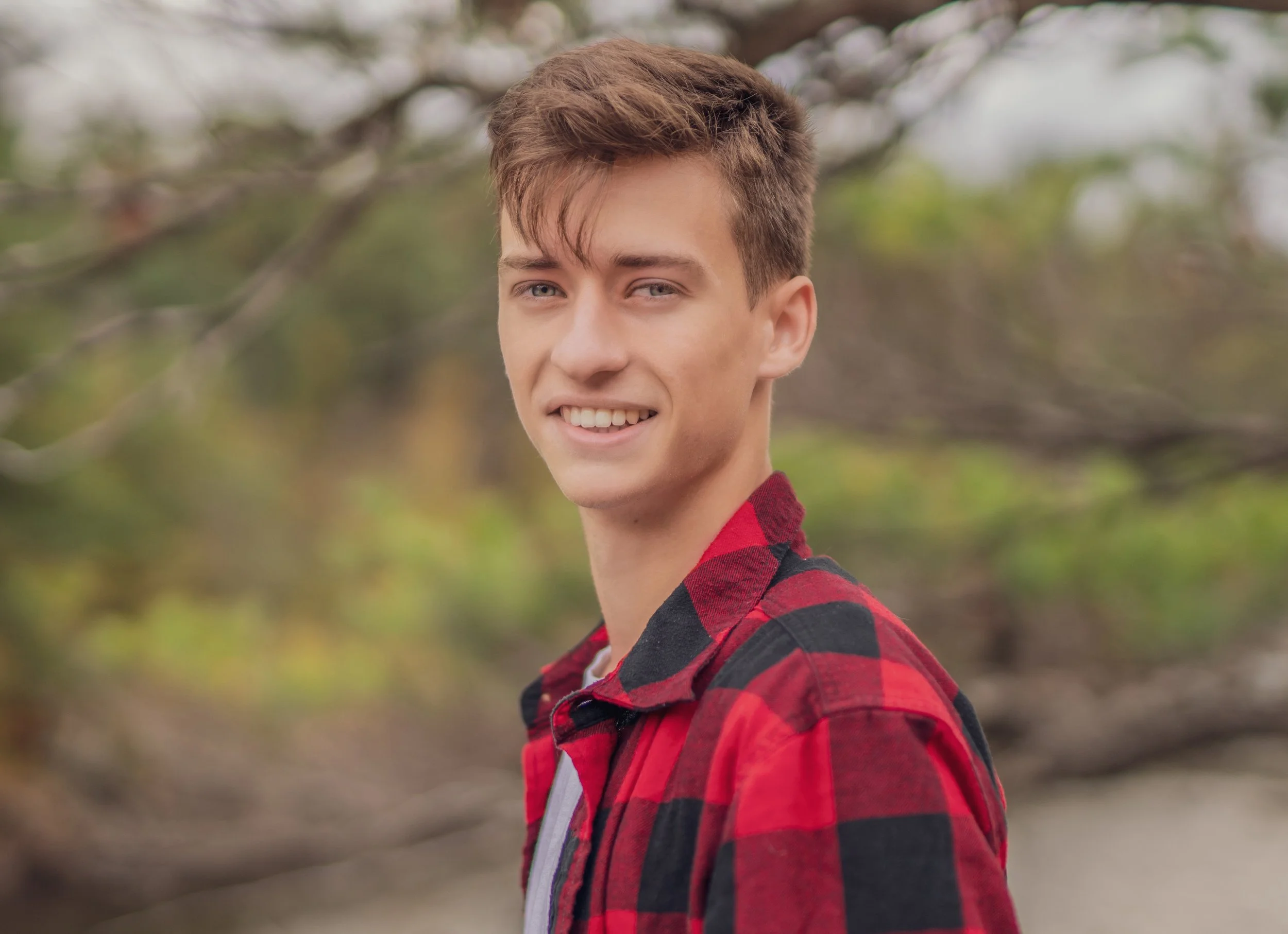 A young man with light brown hair smiling outdoors while wearing a red and black plaid shirt, with blurred greenery in the background.