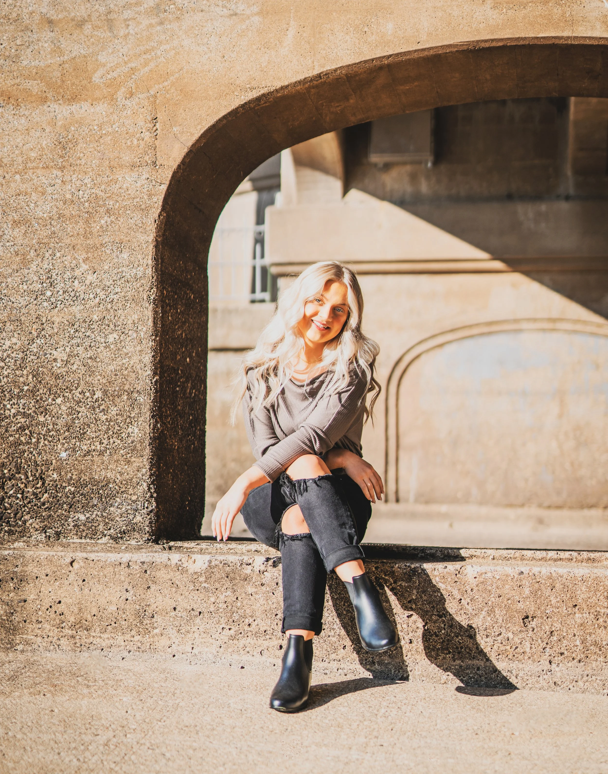 A young woman with blonde hair and a gray sweater sitting on a concrete ledge under an archway in sunlight, smiling at the camera.