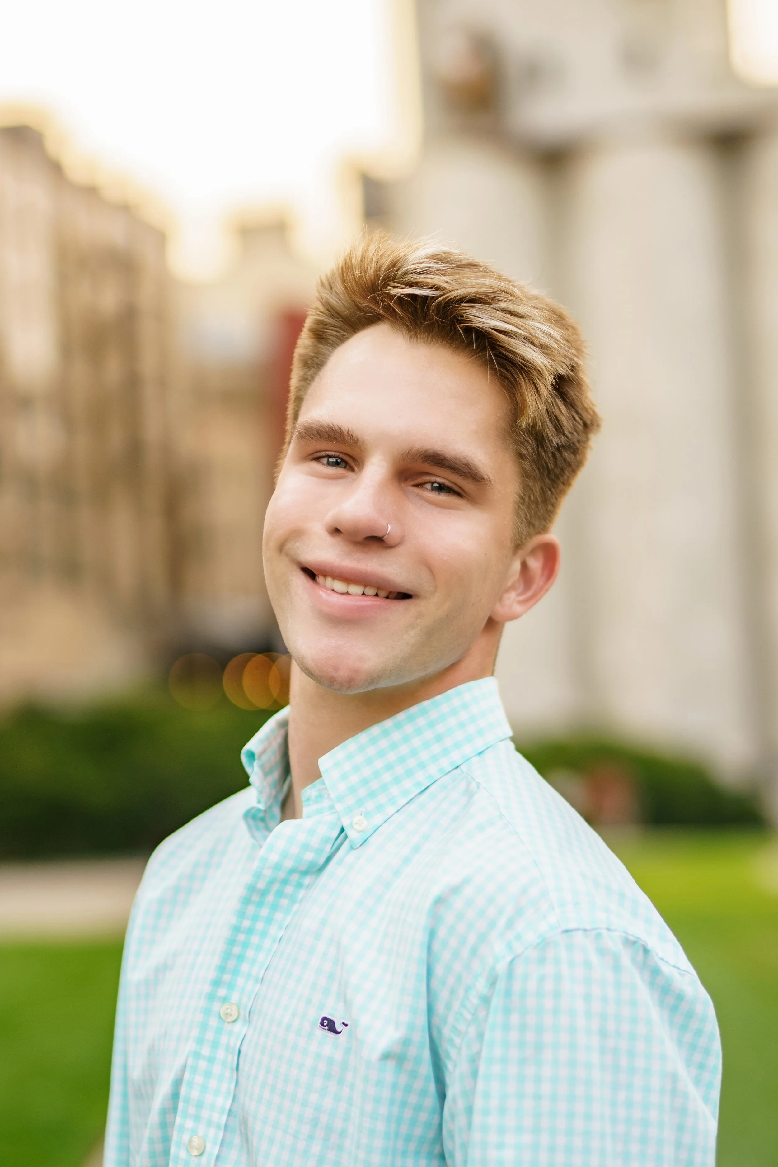 A young man smiling outdoors with buildings and greenery in the background.