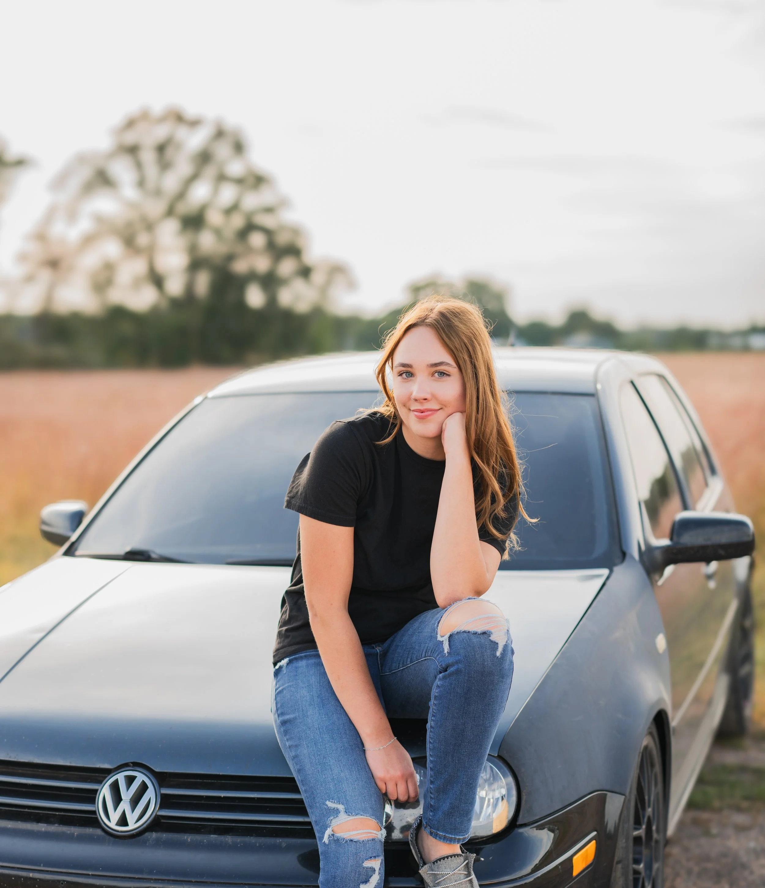 A young woman with red hair wearing a black t-shirt and ripped jeans sitting on the hood of a dark gray Volkswagen car in an open field.