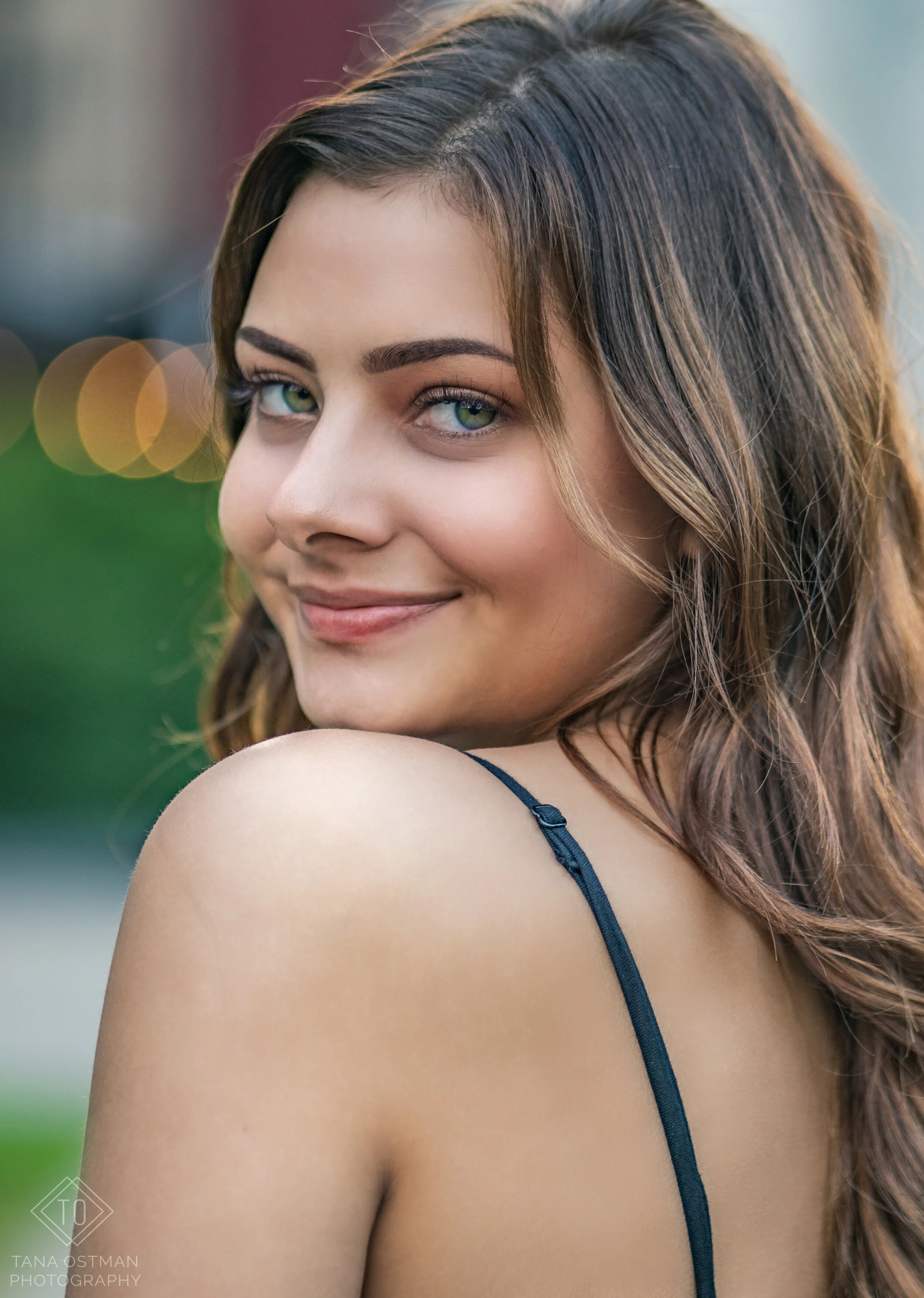 Close-up of a young woman with long, wavy brown hair and bright blue eyes, smiling and looking over her shoulder in an outdoor setting with green blurred background.