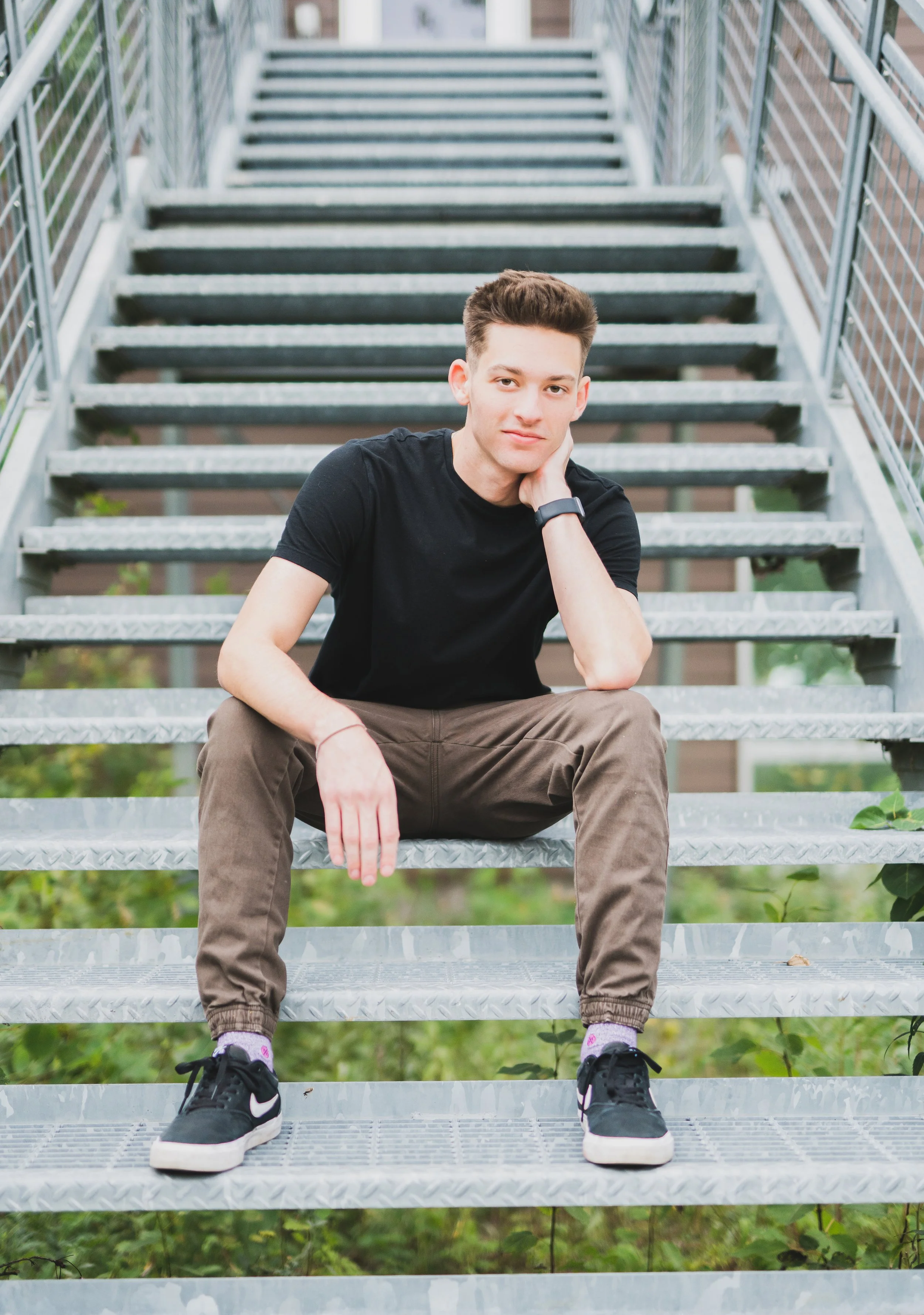 A young man sitting on metal outdoor stairs, resting his head on his hand, wearing a black t-shirt, brown pants, black sneakers, and a wristwatch, with greenery visible beneath the stairs.