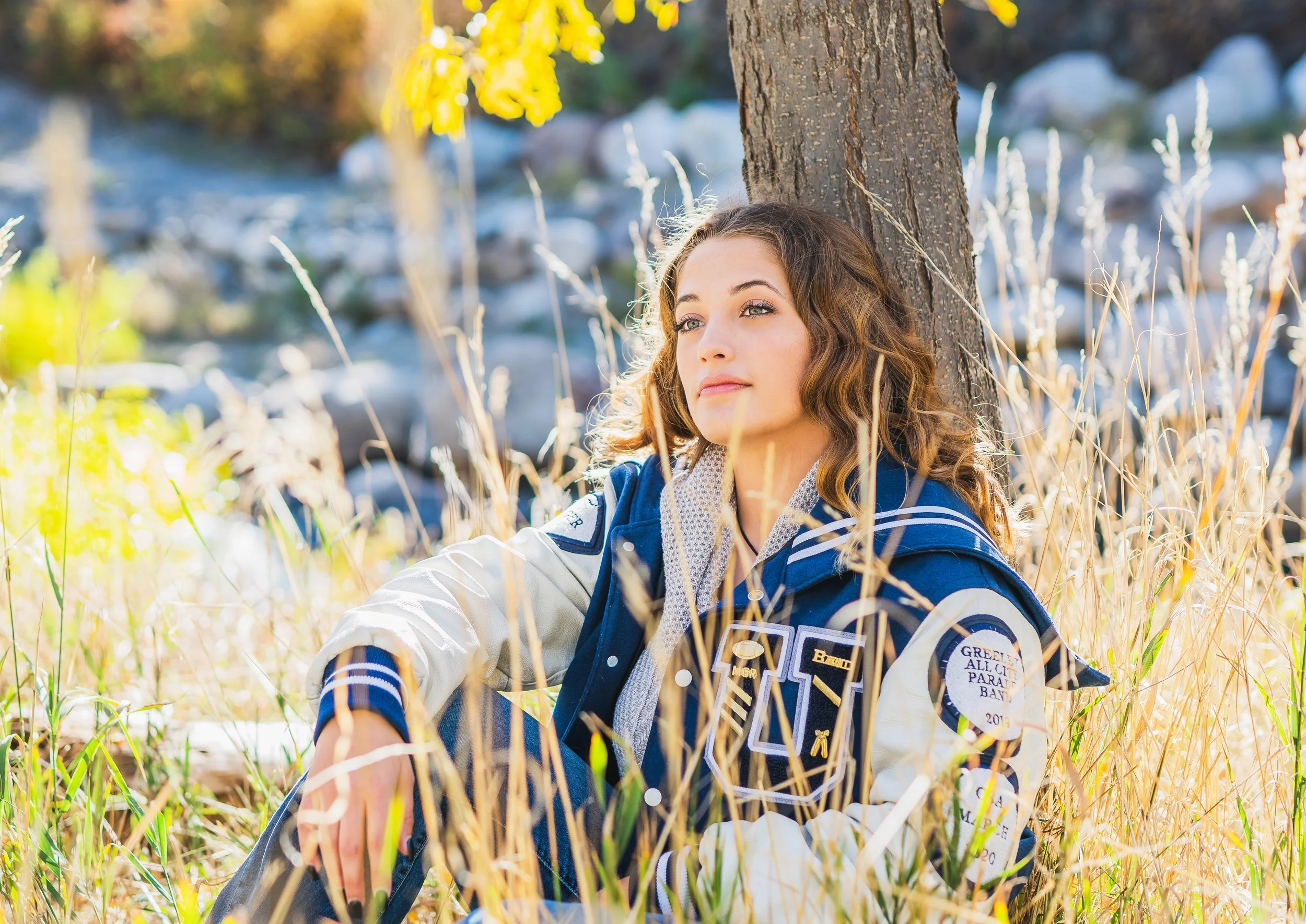 A young woman sitting in a grassy field next to a tree, with a rocky background, wearing a Girl Scout uniform and looking pensively into the distance.