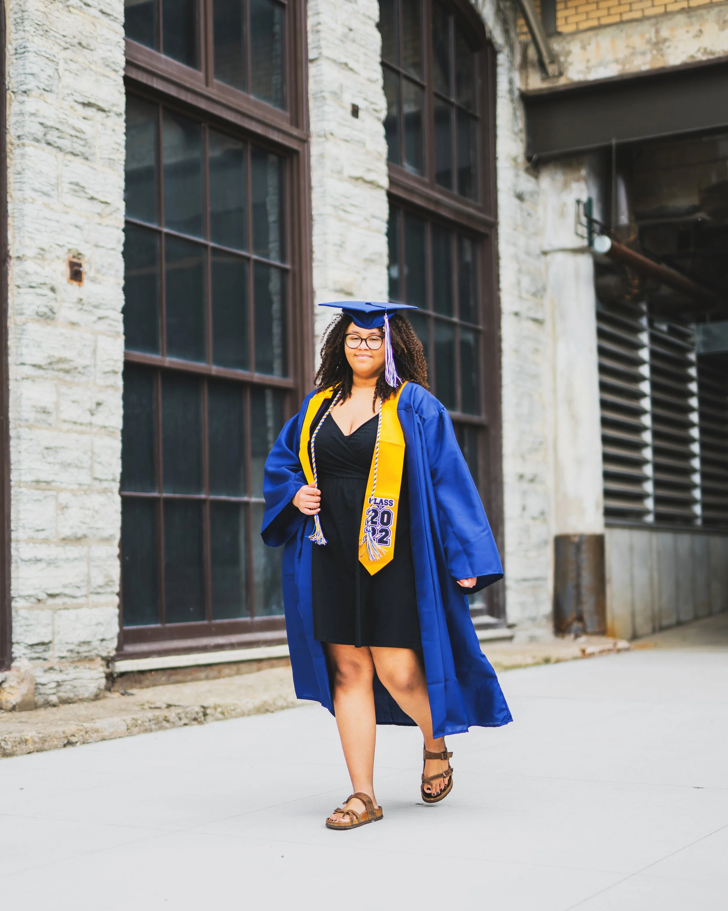 A woman wearing a blue graduation gown and cap with a yellow sash and a Class of 2022 stole, walking outdoors in front of a building with large windows.