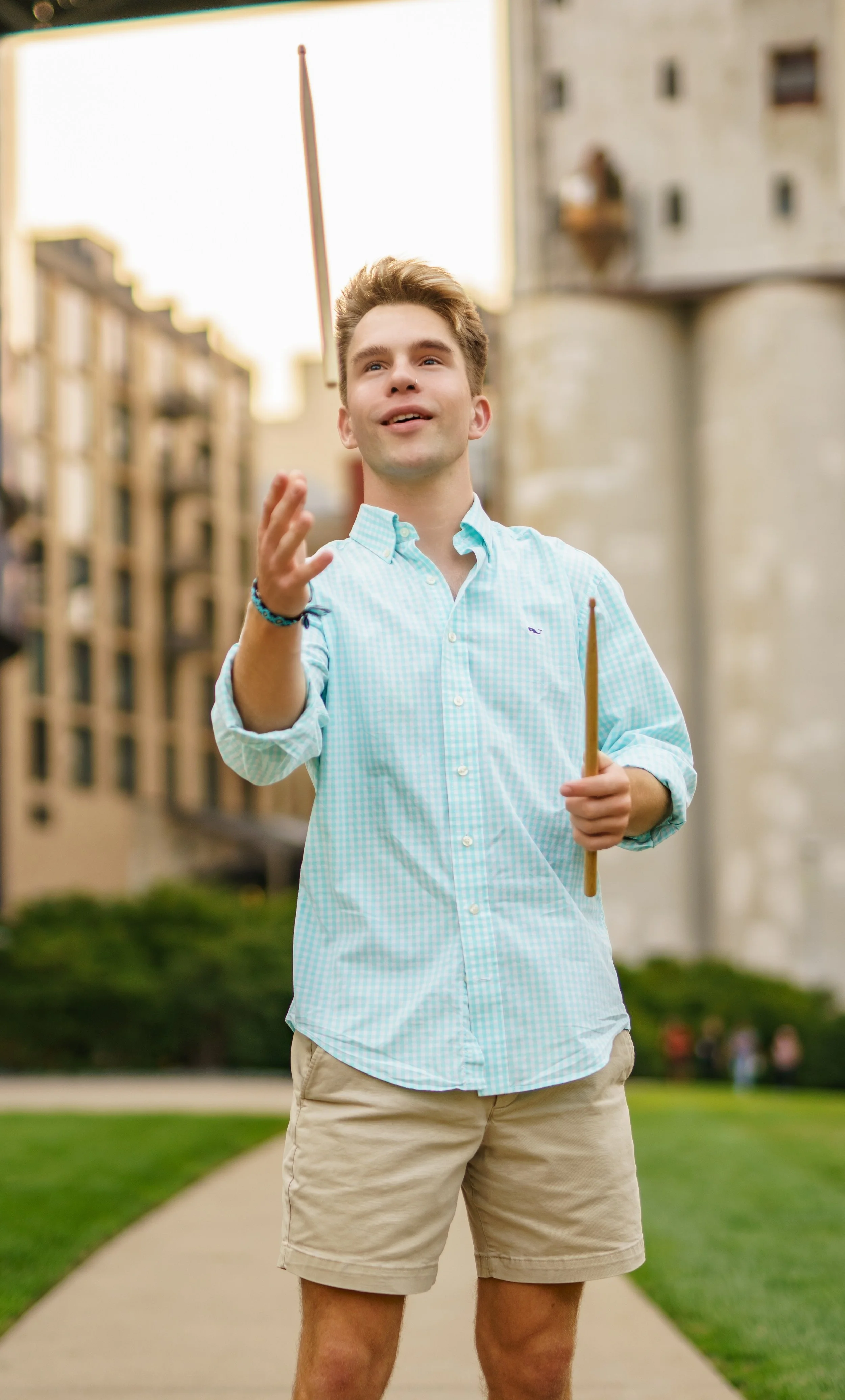A young man in a checkered blue shirt and khaki shorts holding a baton, standing outdoors on a sidewalk with buildings and greenery in the background.
