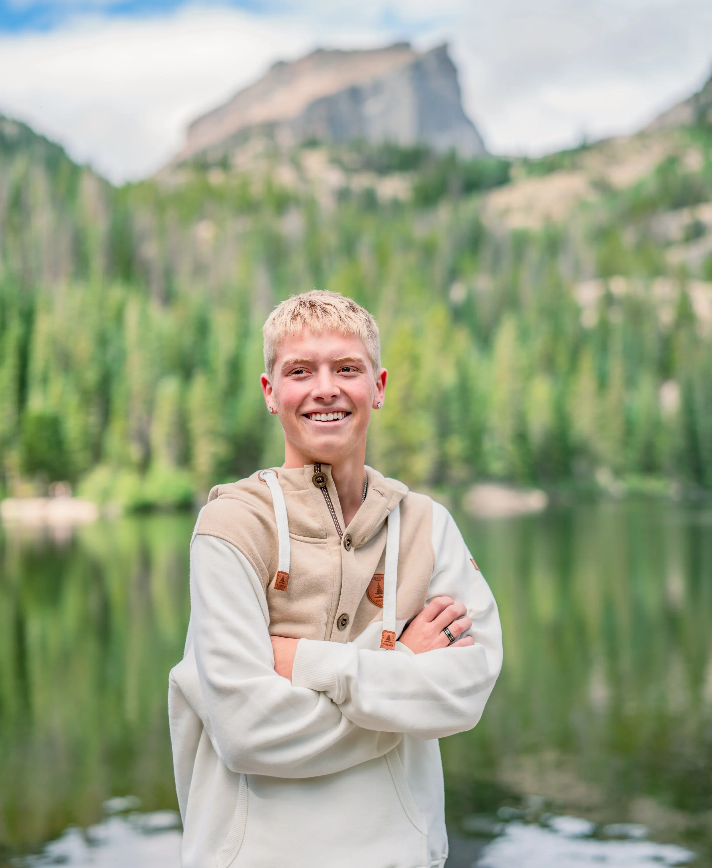 A smiling young person standing outdoors in front of a forested mountain landscape, with arms crossed and a lake behind them.