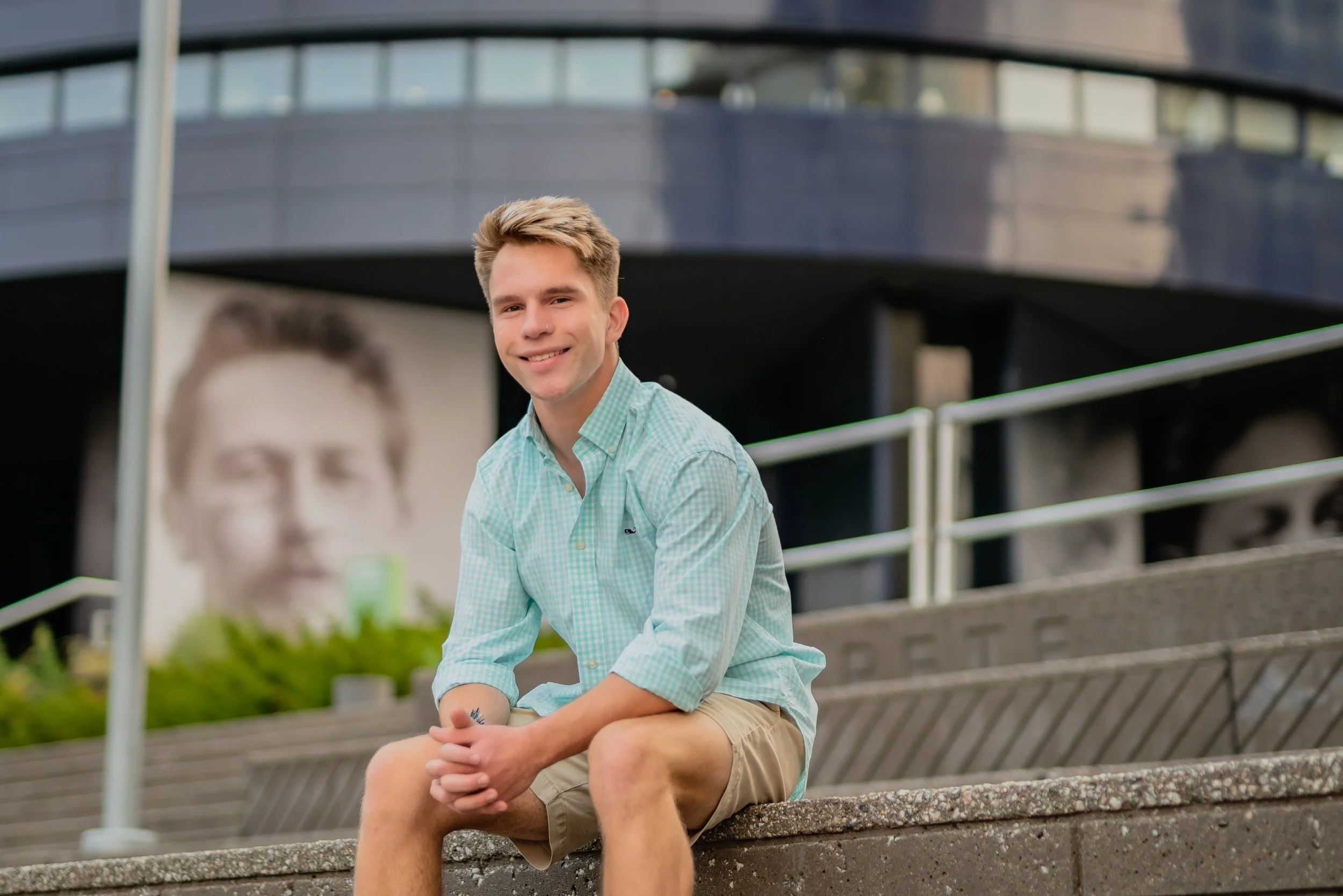 A young man in a light green checkered shirt and beige shorts sitting on a concrete ledge outdoors, smiling at the camera. In the background, there is a black and white portrait of a person and a modern building with reflective windows.