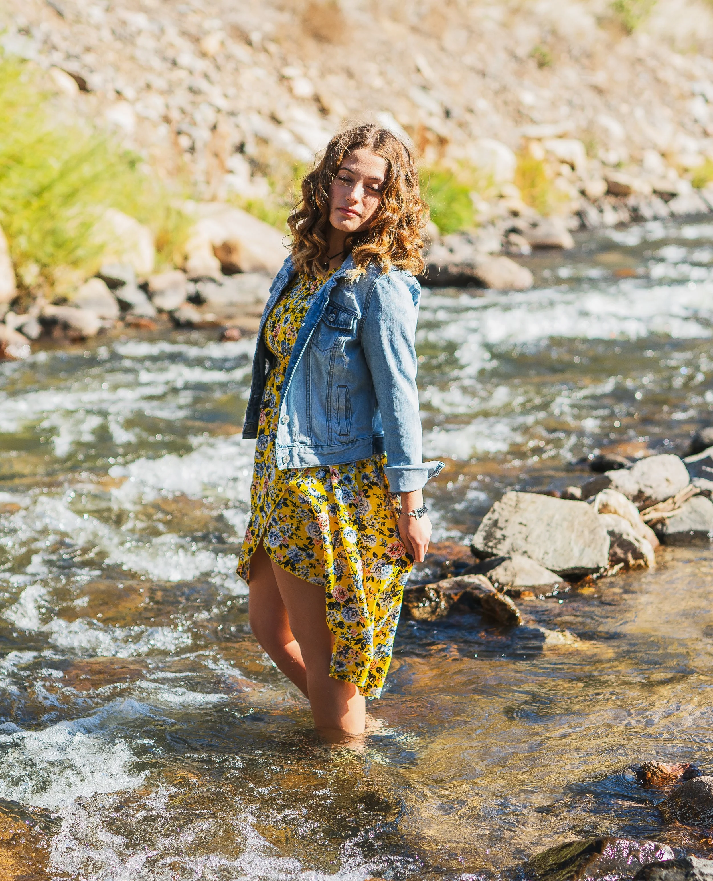A woman in a yellow floral dress and denim jacket standing in a rocky stream with water reaching her knees, surrounded by nature.
