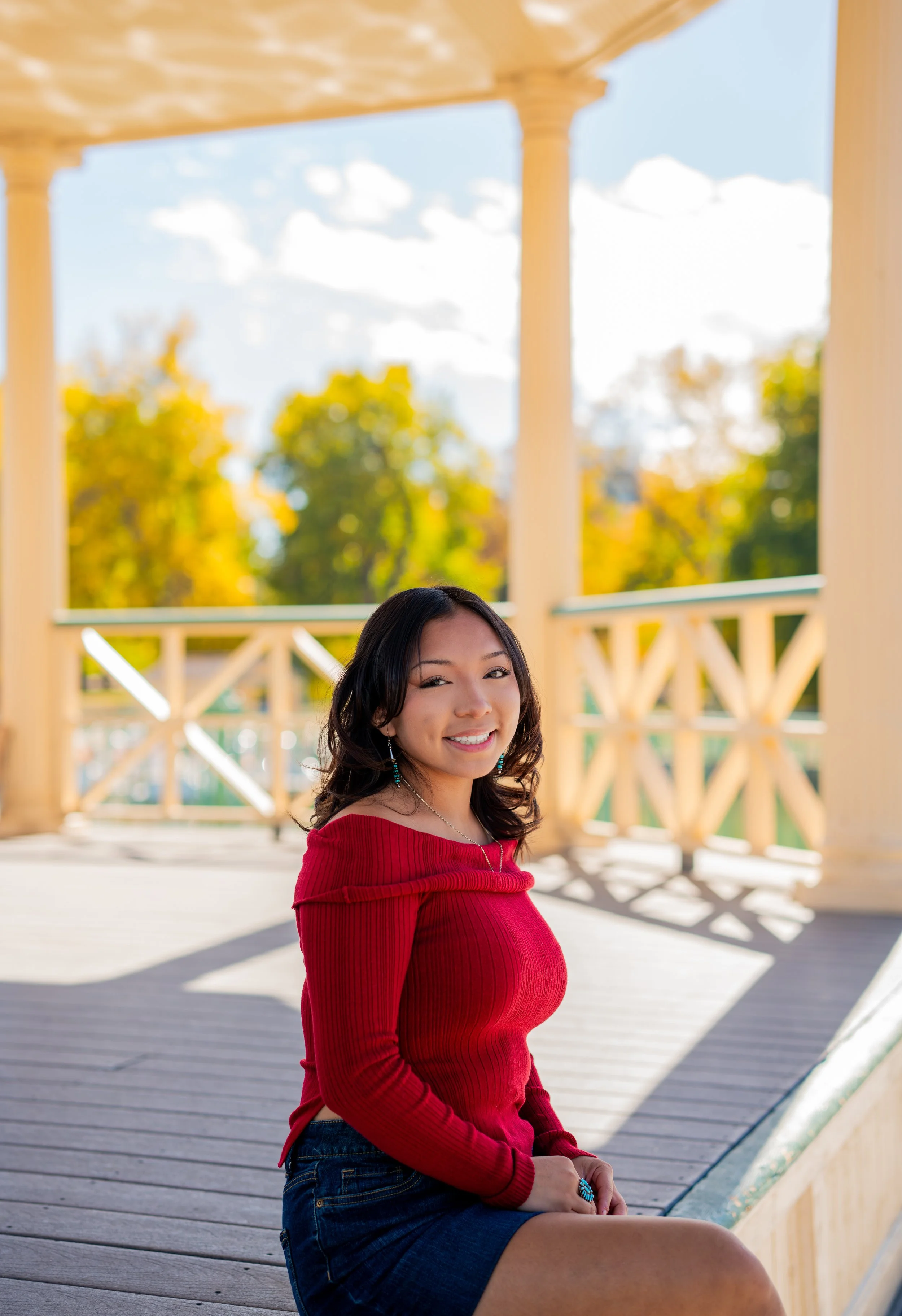 A woman with long dark hair wearing a red off-the-shoulder top and denim skirt, sitting on a wooden porch with yellow columns and railing, smiling at the camera with trees and sky in the background.