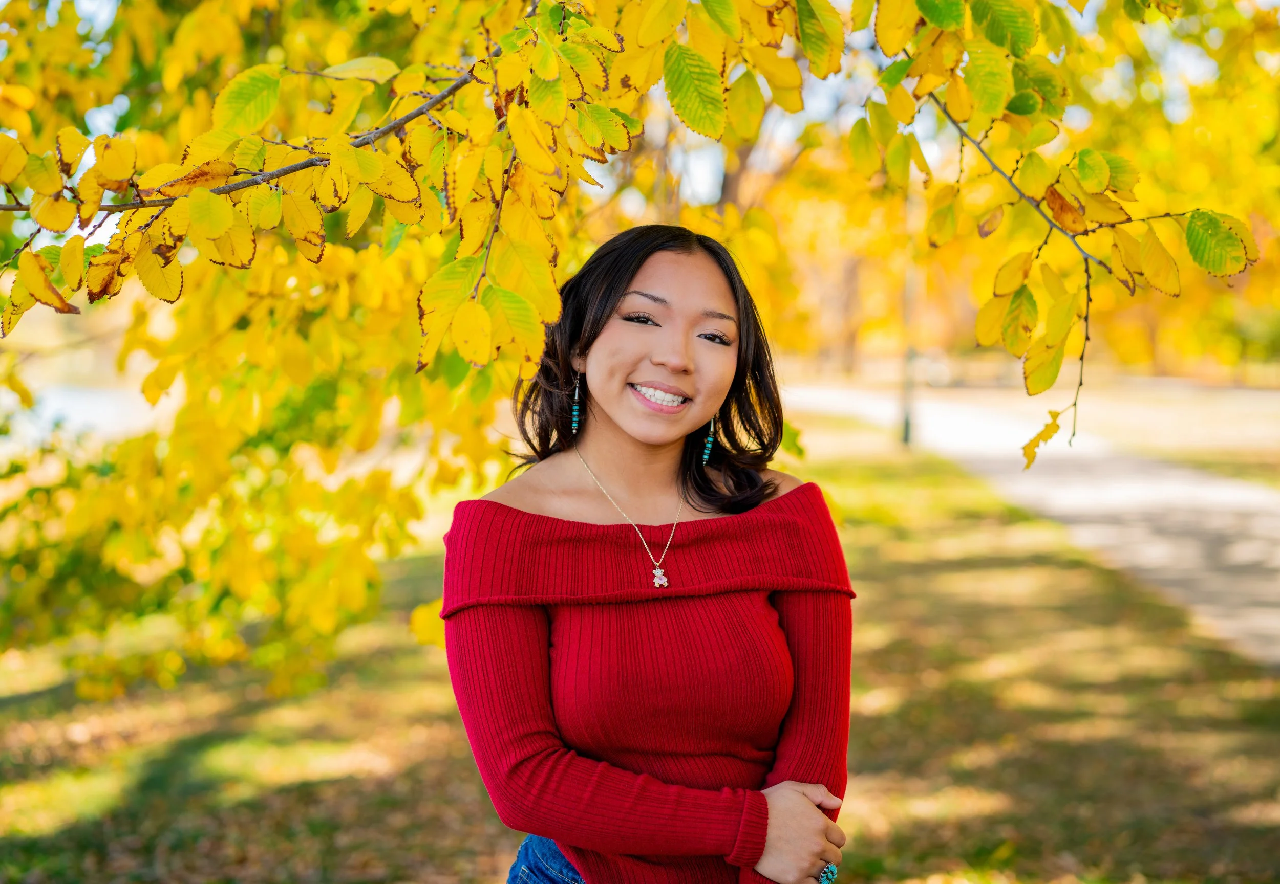 A young woman with long dark hair smiling in autumn outdoors, wearing a red off-the-shoulder sweater, earrings, and necklace, standing under yellow and green tree foliage.