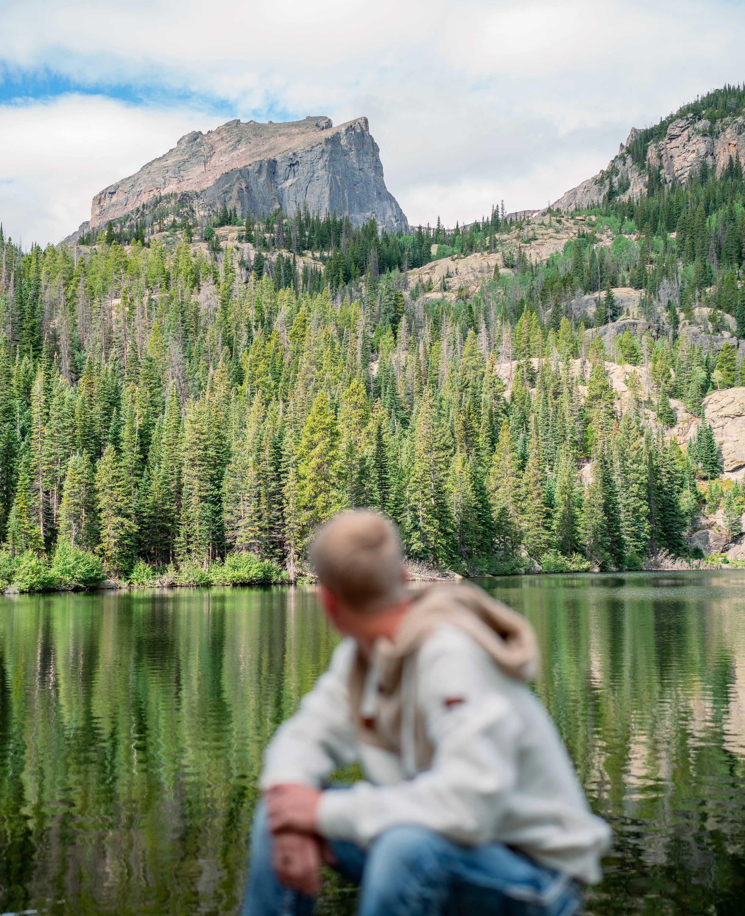 A person with a blurred face sitting by a lake surrounded by a dense forest with mountains in the background.