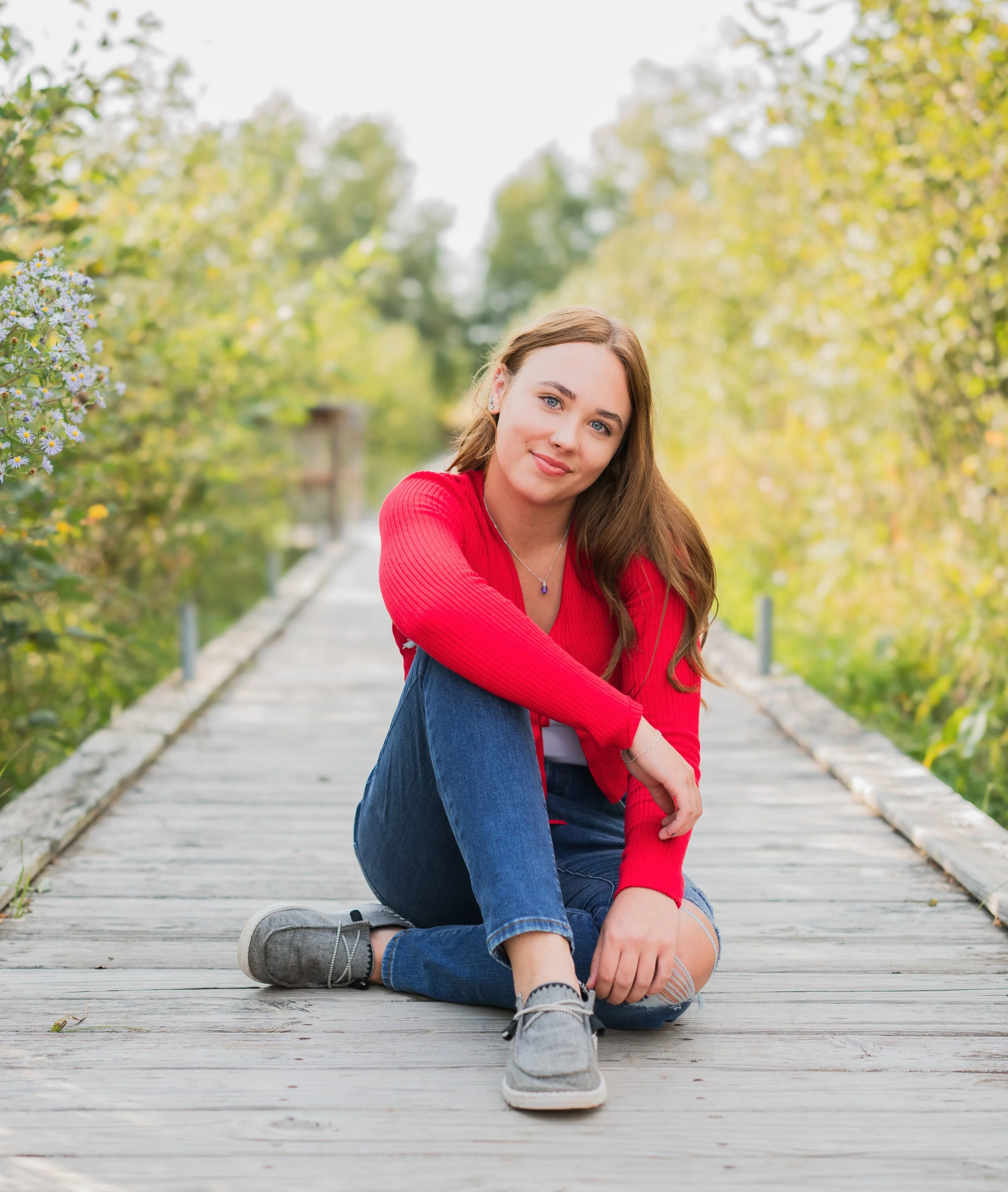 A young women with long brown hair, wearing a red long-sleeve shirt, ripped jeans, and gray shoes, sitting on a wooden bridge in a natural outdoor setting with green trees and yellow foliage.