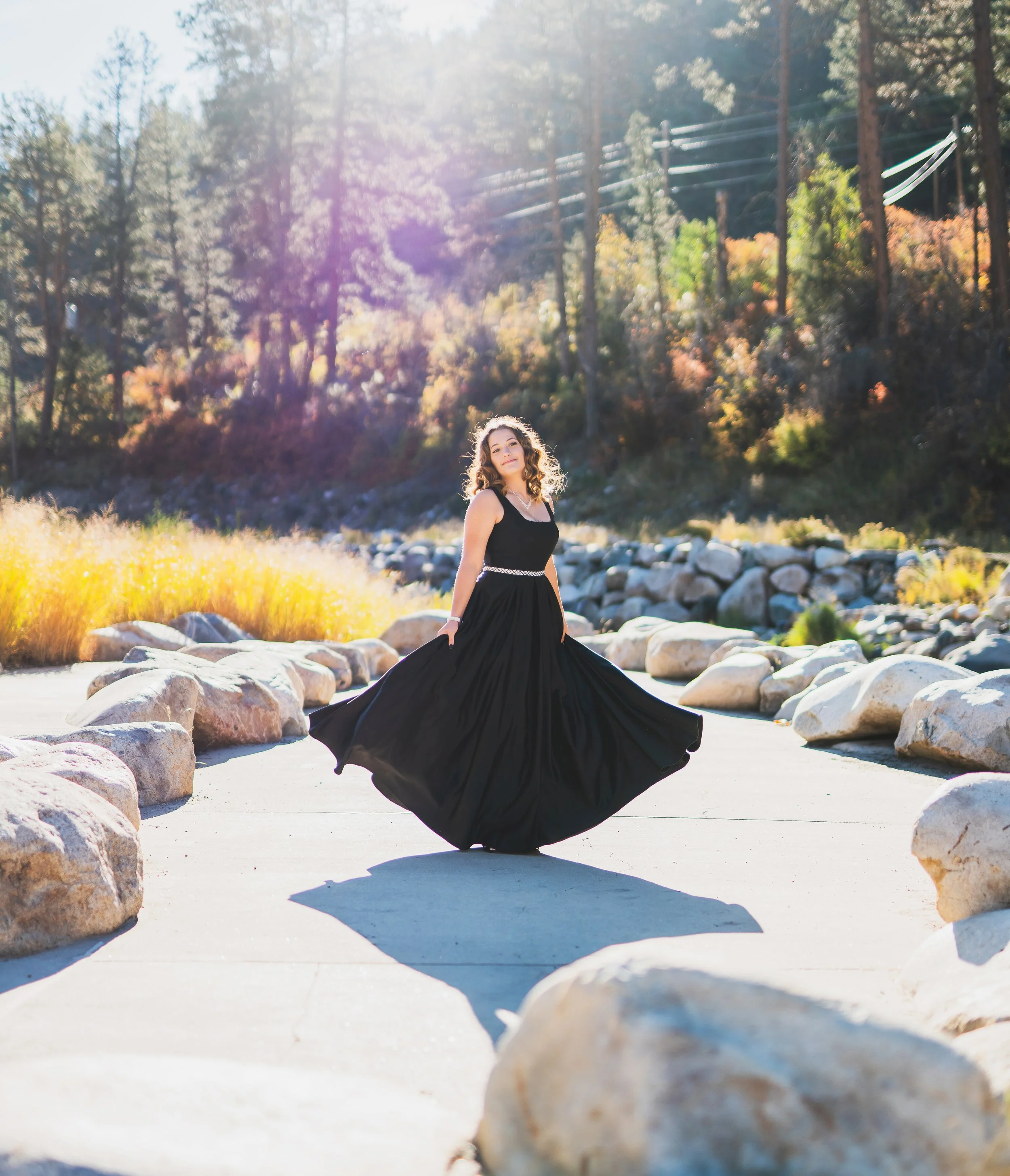 A woman in a flowing black dress standing on a path lined with large rocks, with trees and sunlight in the background.